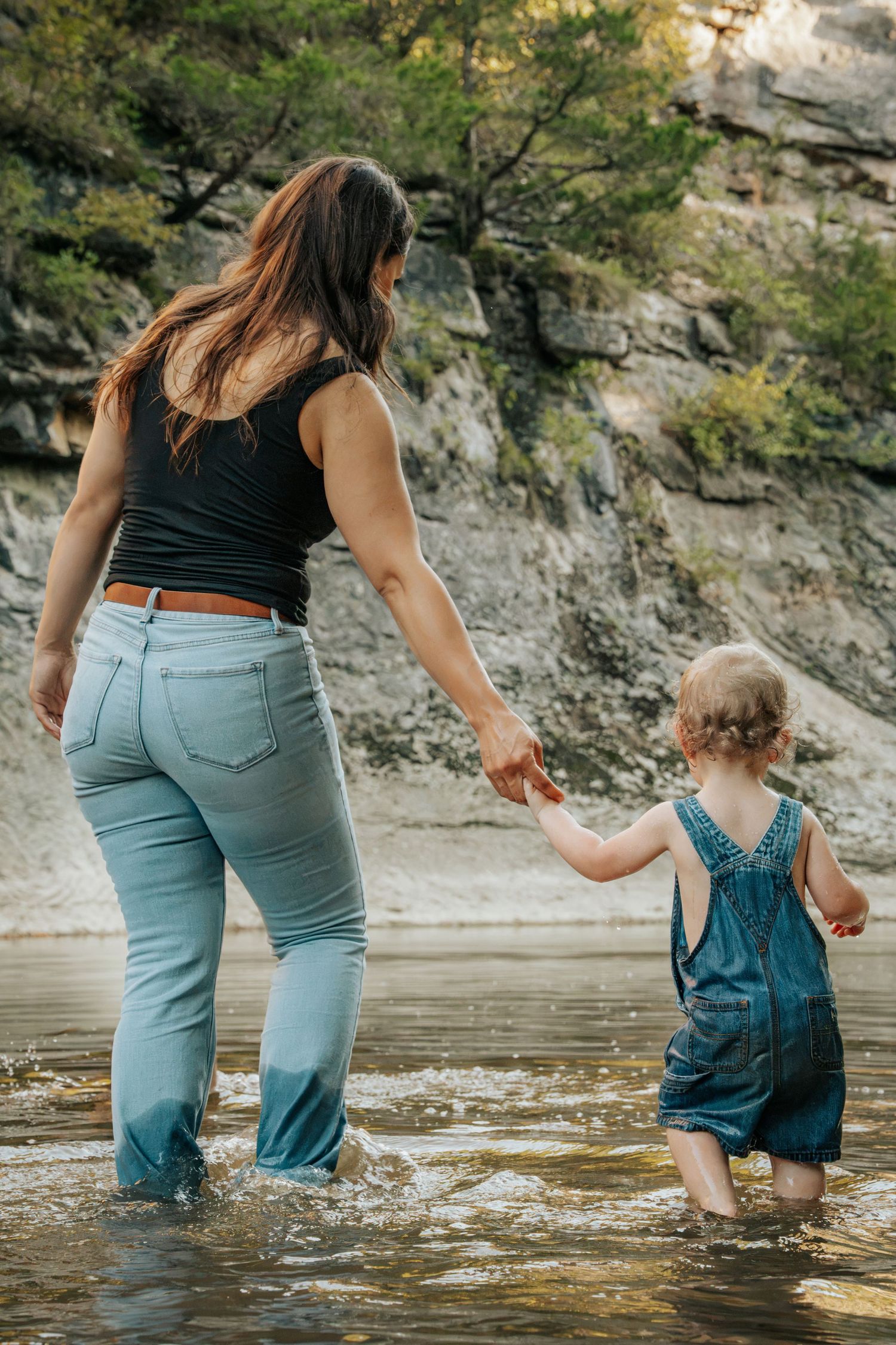 A mother and toddler girl in denim overalls hold hands while wading through a shallow rocky river.