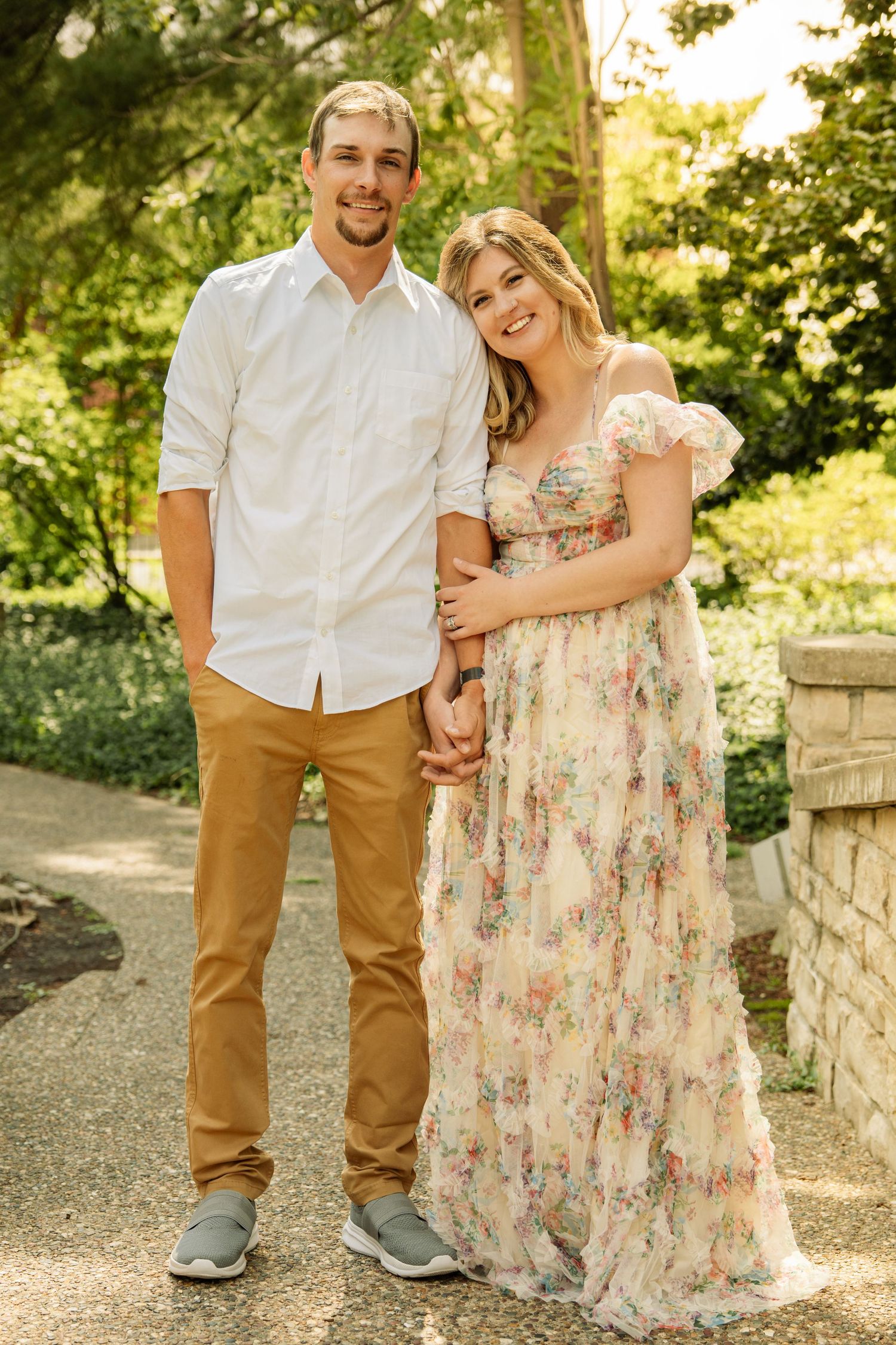 A smiling couple poses in a lush garden, man in white shirt and tan pants, woman in floral maxi dress.