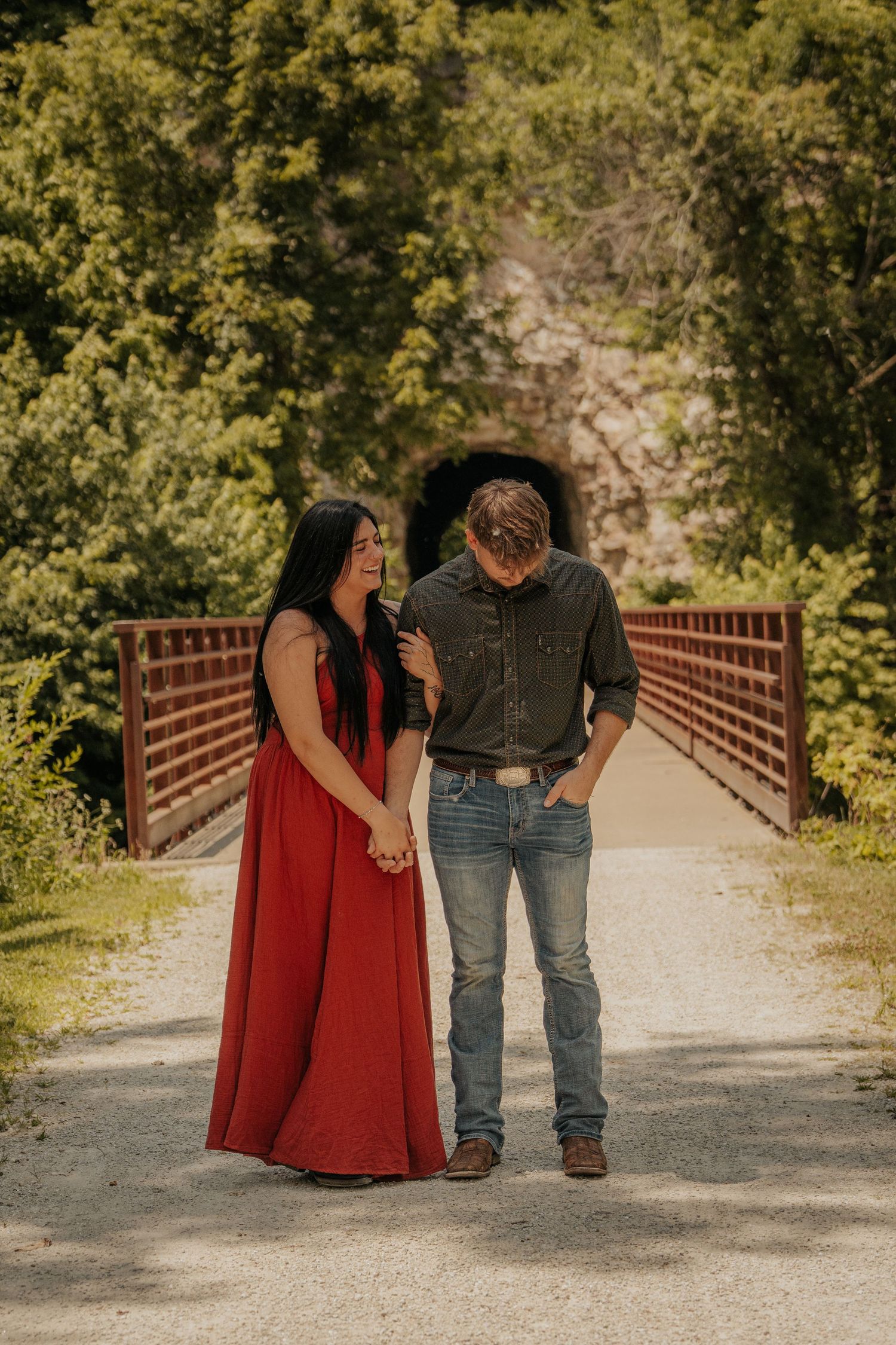 Couple holding hands on a bridge path, woman in red maxi dress, man in jeans, with a stone tunnel and green trees behind.
