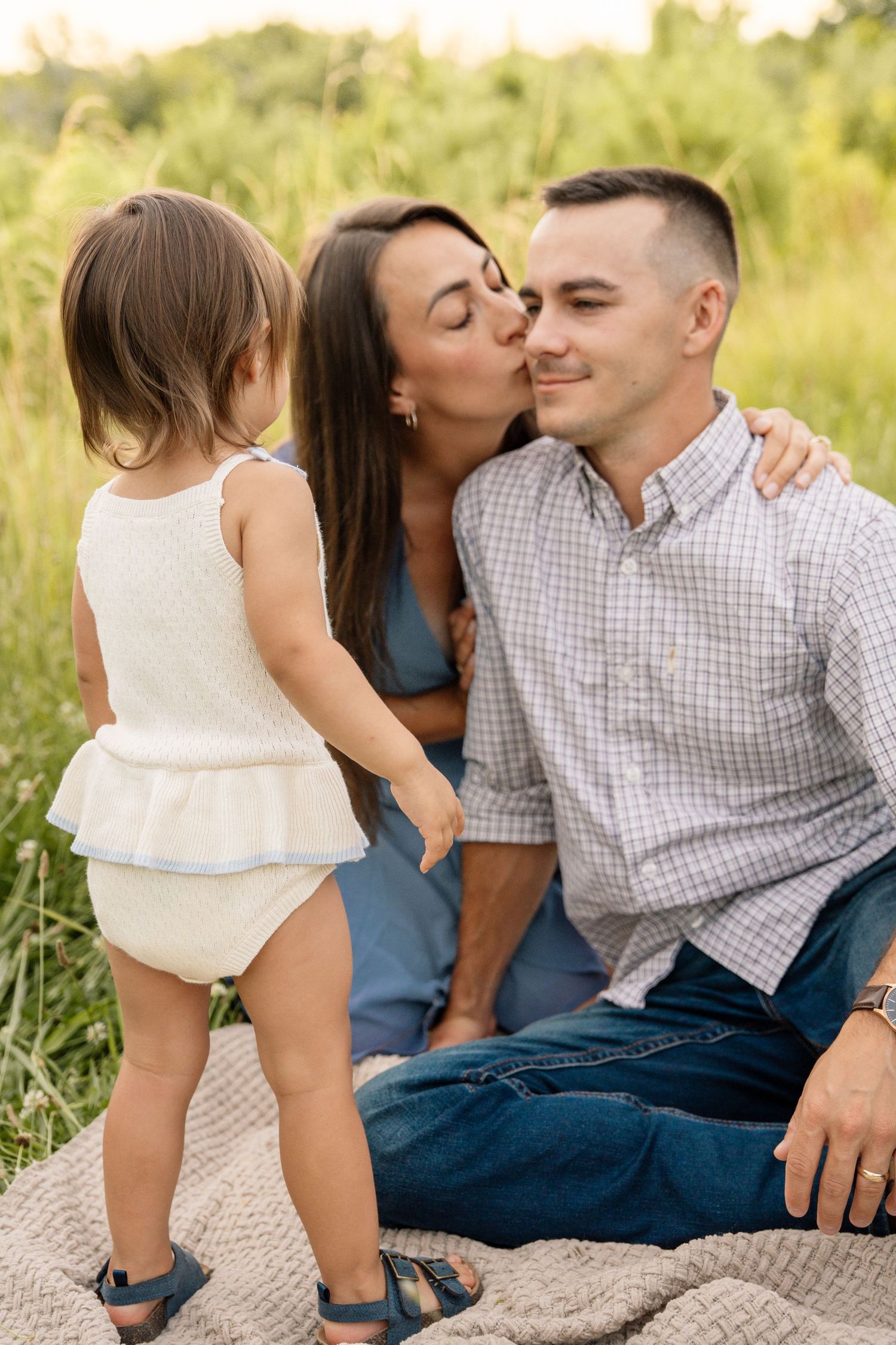 Young family sitting on blanket in green field, mother kissing fathers cheek while toddler stands nearby.
