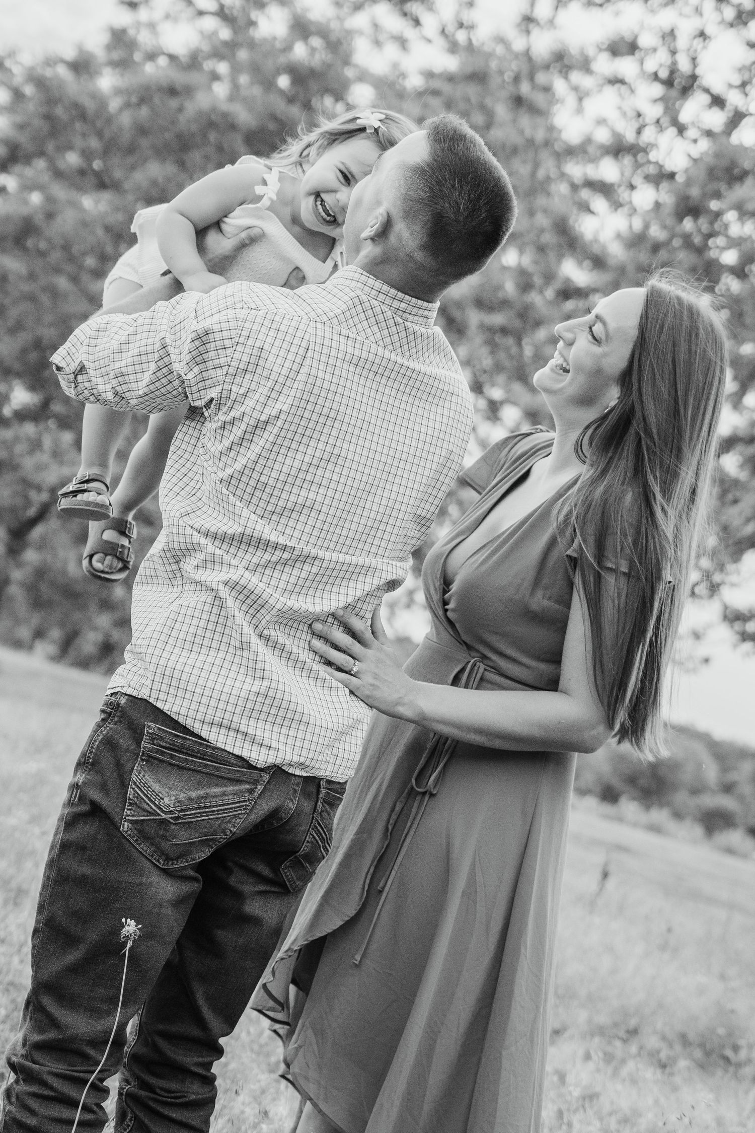 Black and white photo of a happy family with father lifting laughing toddler while mother smiles in a park.