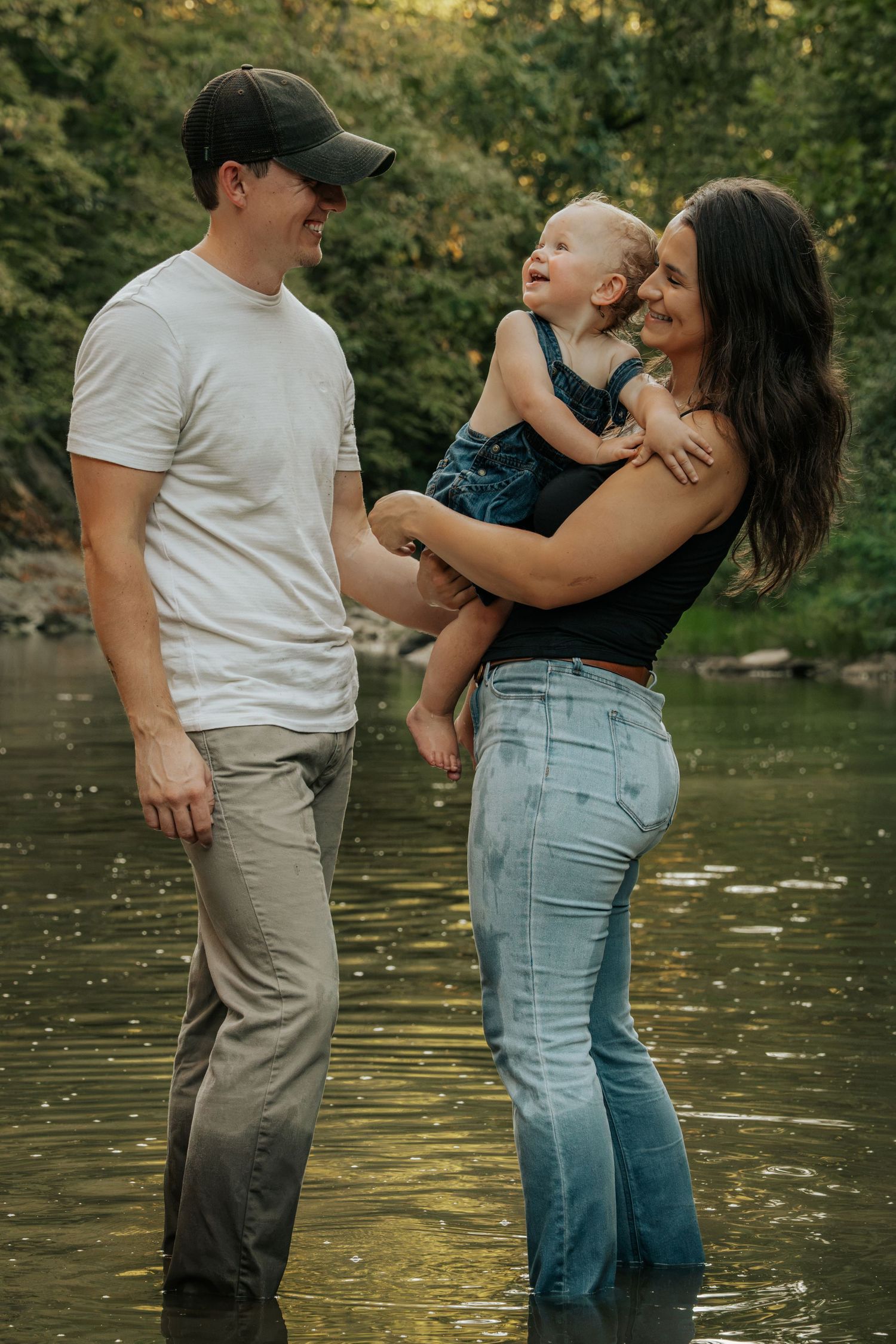A happy young family stands in a shallow river, with mom holding a smiling toddler while dad looks on lovingly.