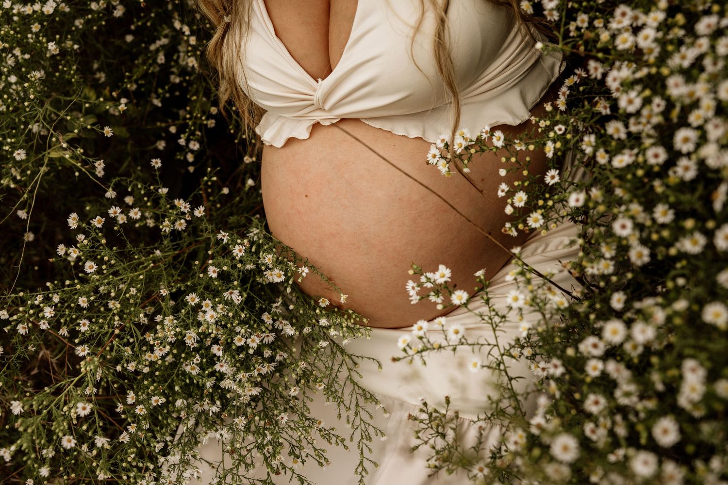 Close-up of a pregnant belly surrounded by delicate white wildflowers, wearing a white dress outdoors.