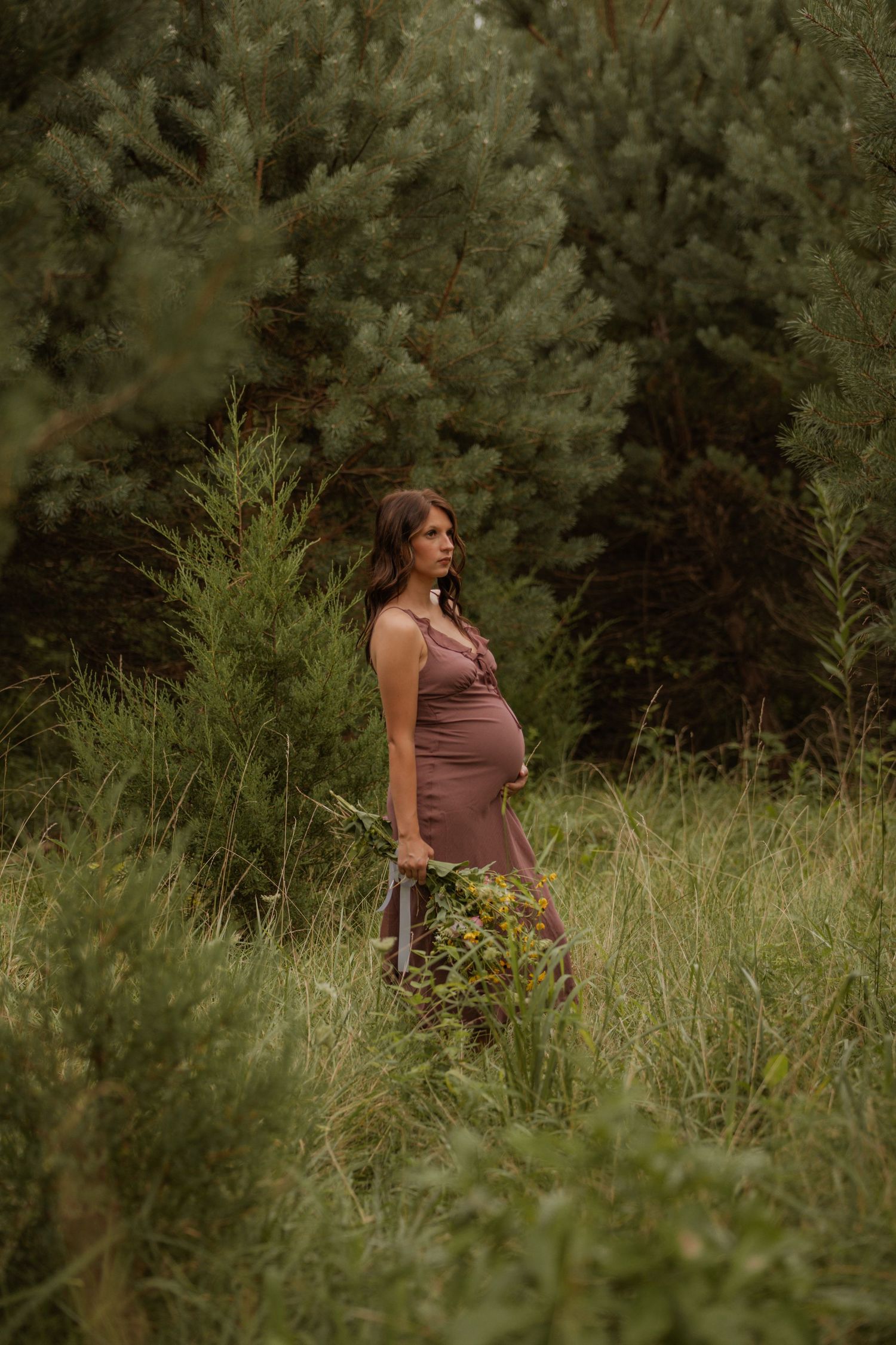Pregnant woman in brown dress holding wildflowers stands in lush green forest meadow.
