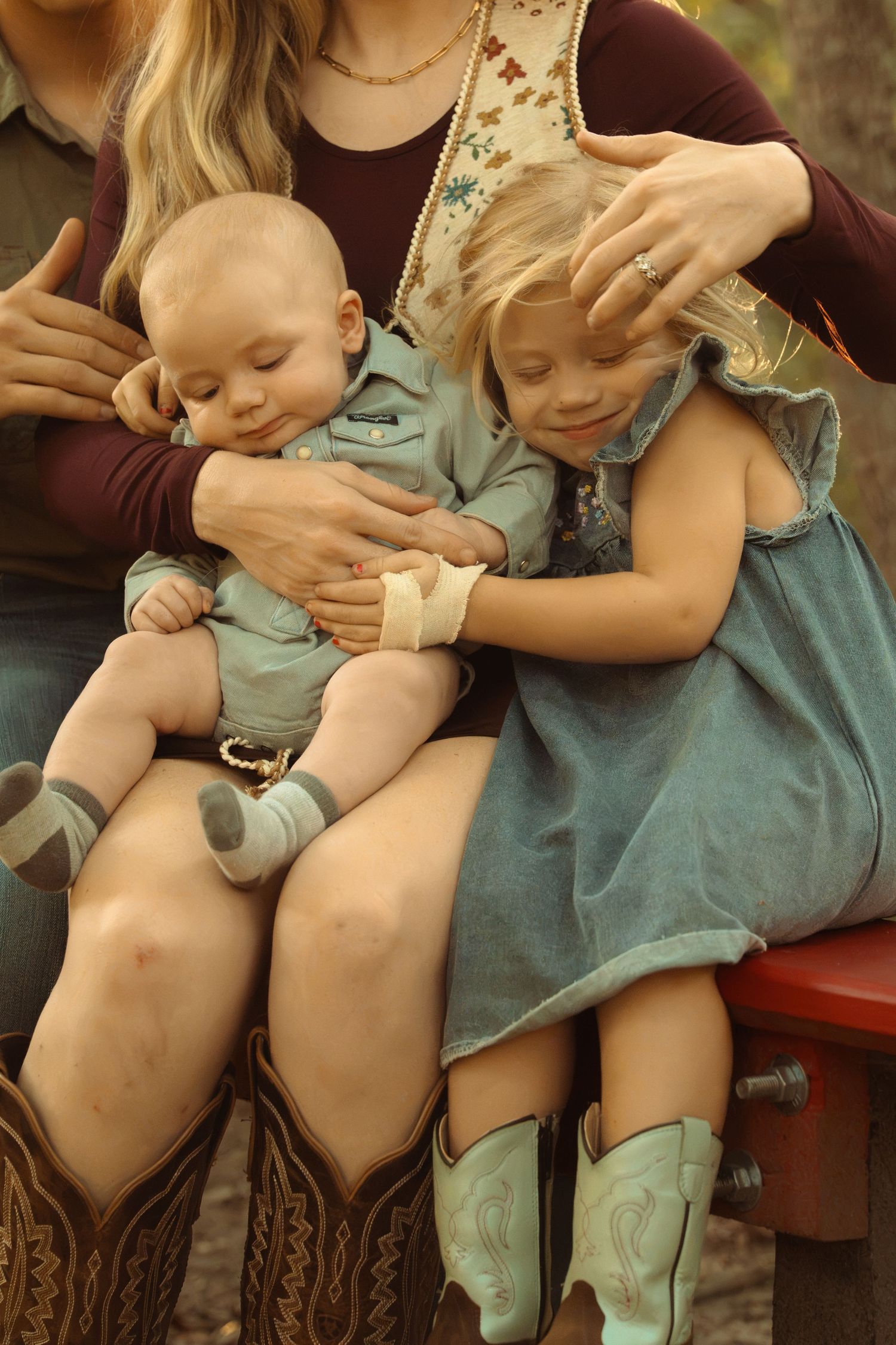 A young girl in a denim dress and teal cowboy boots holds a sleeping baby on a woman's lap outdoors.