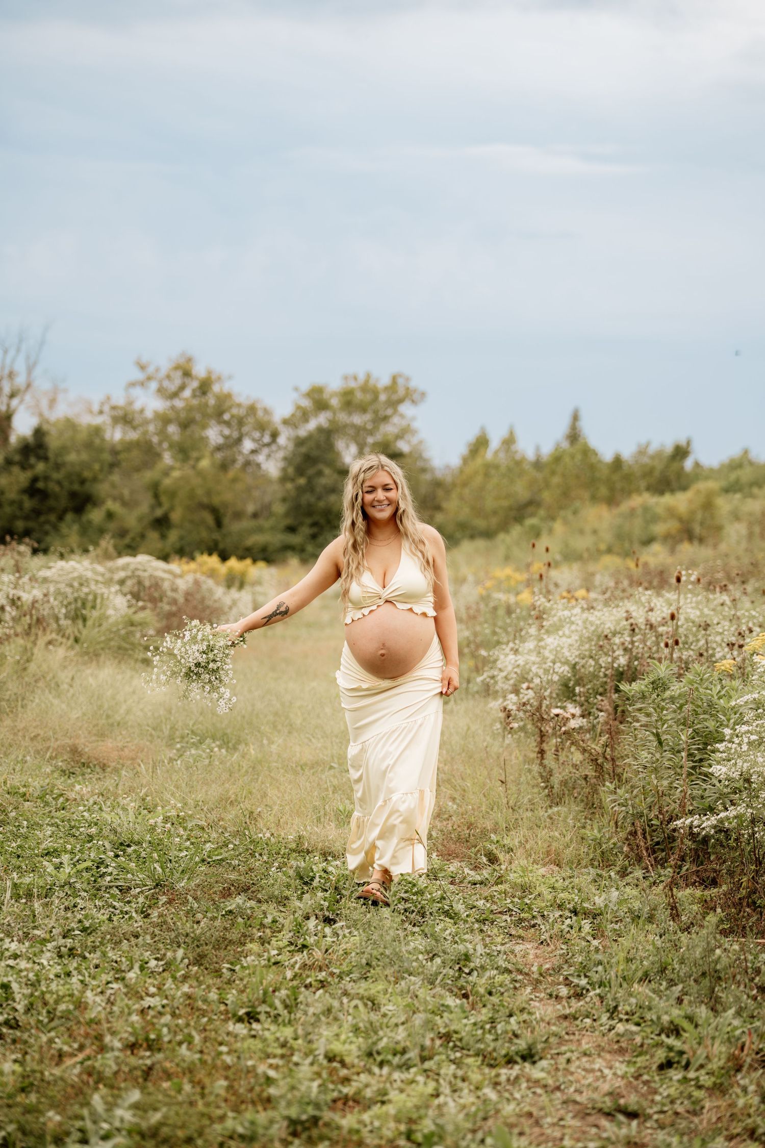 Smiling pregnant woman in cream two-piece maternity outfit walking through wildflower field holding small bouquet.