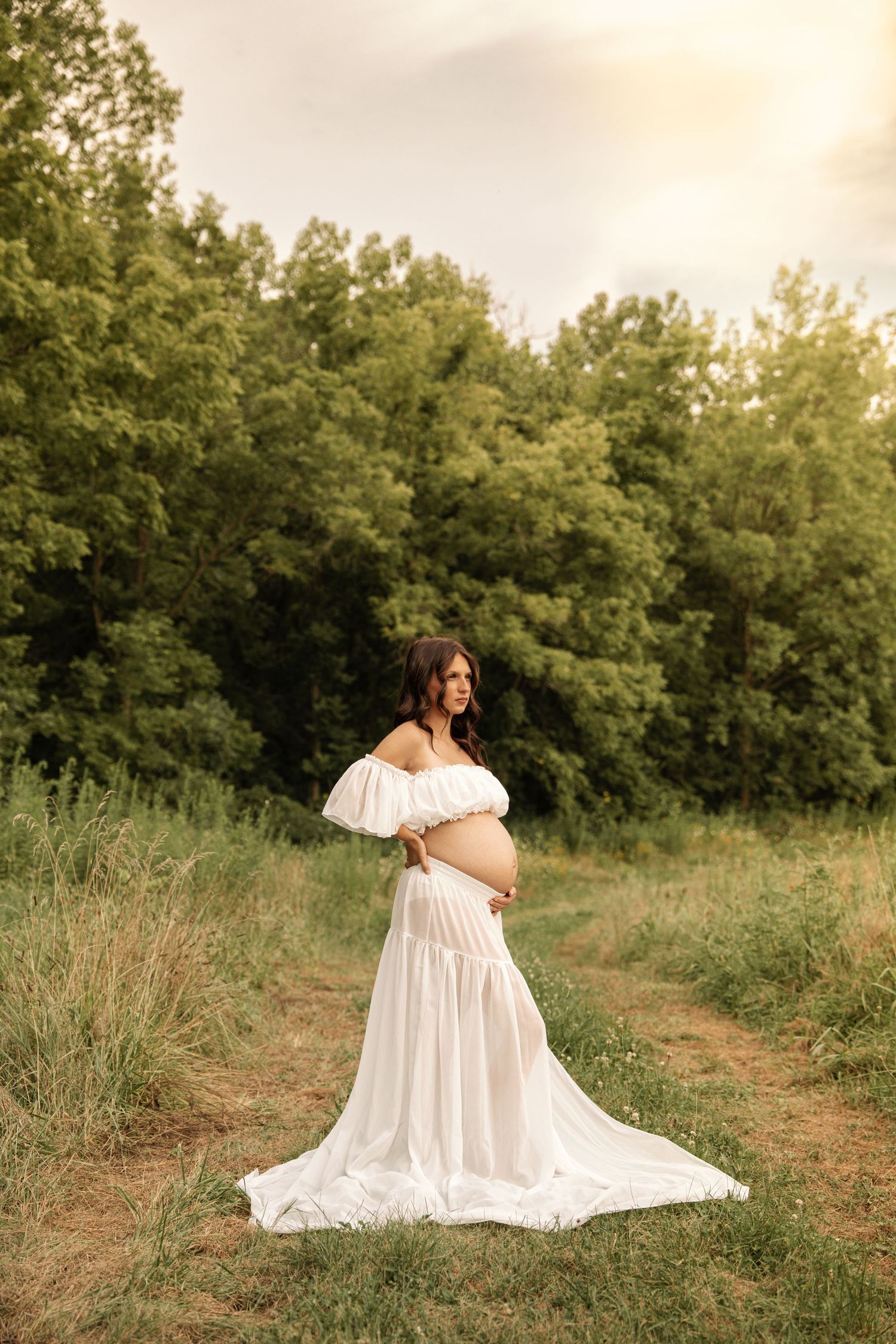 Pregnant woman in white off-shoulder gown cradling baby bump outdoors in lush green field at golden hour.