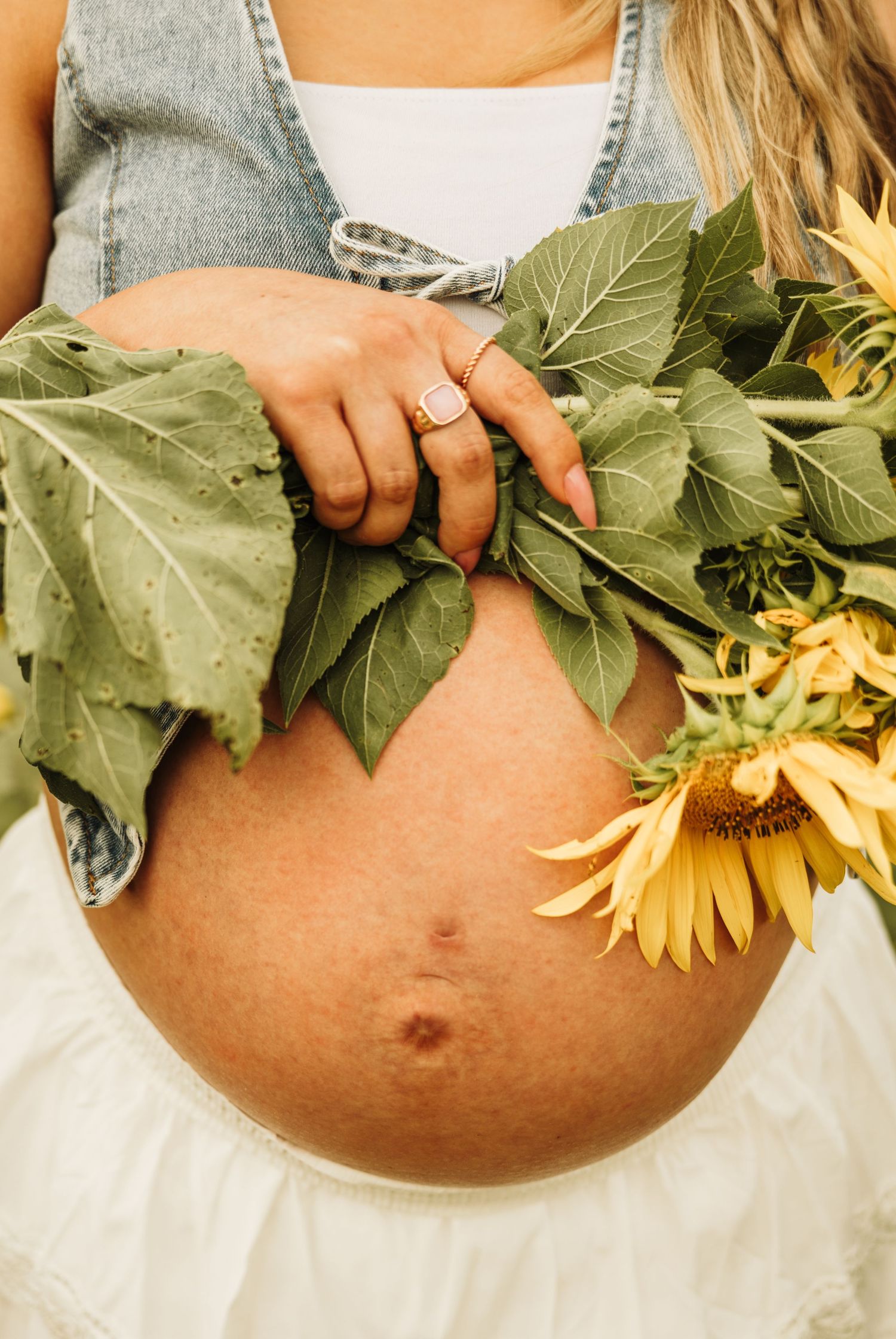 Pregnant woman holding sunflowers against her bare baby bump outdoors, wearing a denim vest and white skirt.