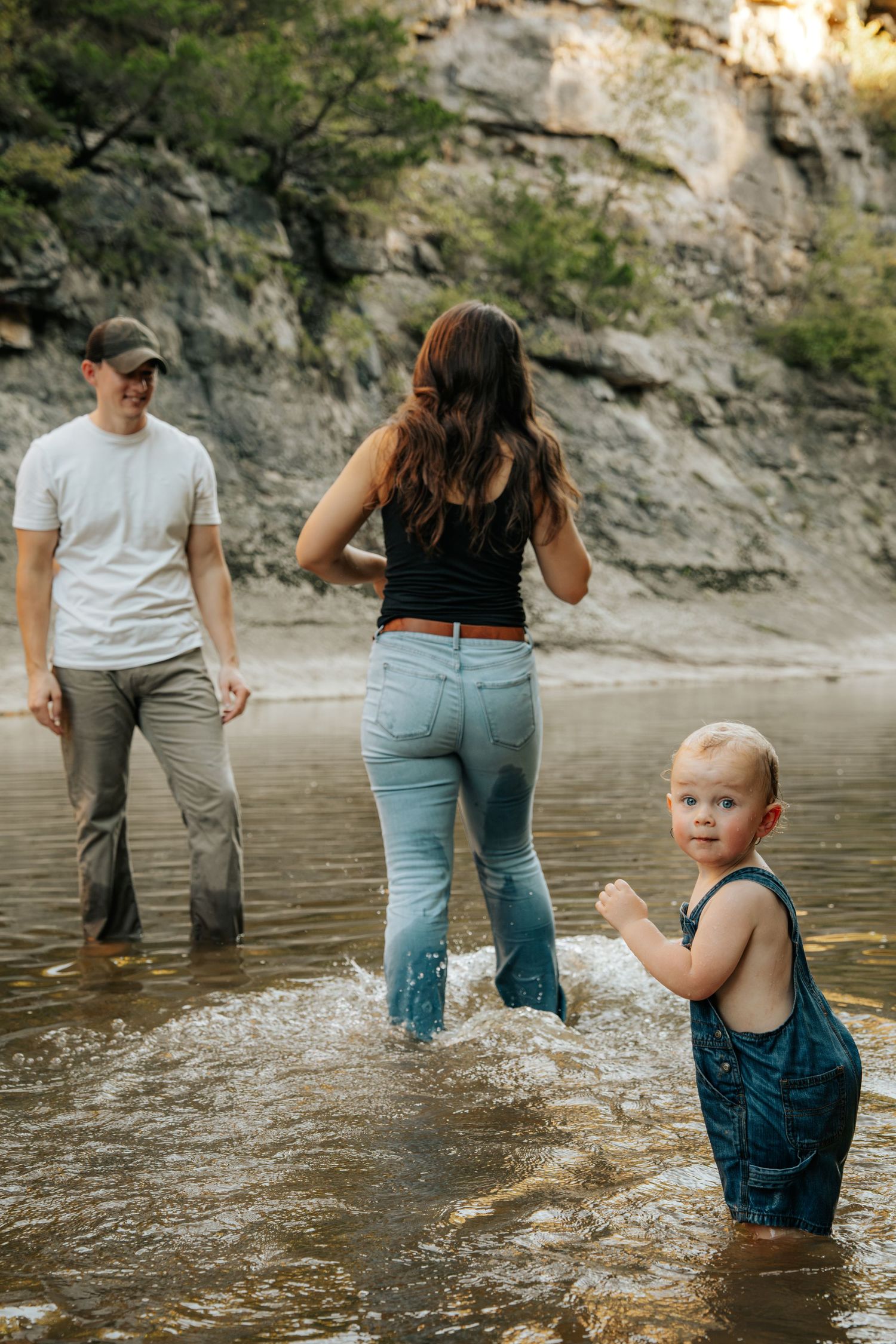 A young family with a blonde toddler in overalls splashes together in a shallow river near rocky cliffs.