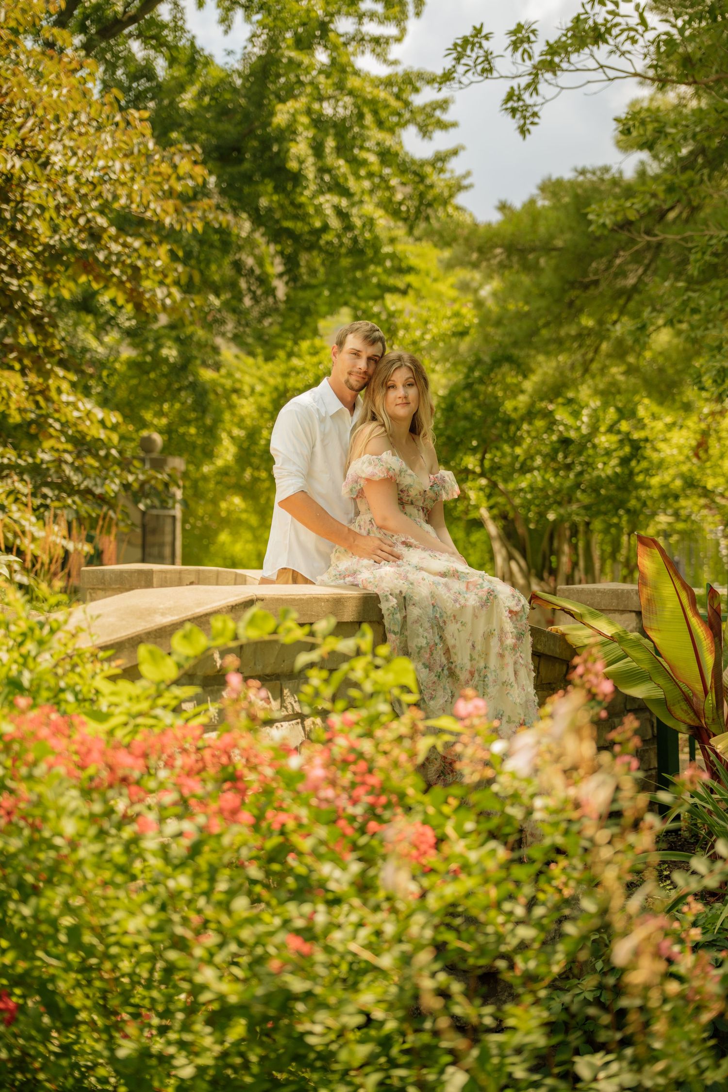 Romantic couple sitting together on a stone bridge surrounded by lush green trees and colorful flowers in a garden.