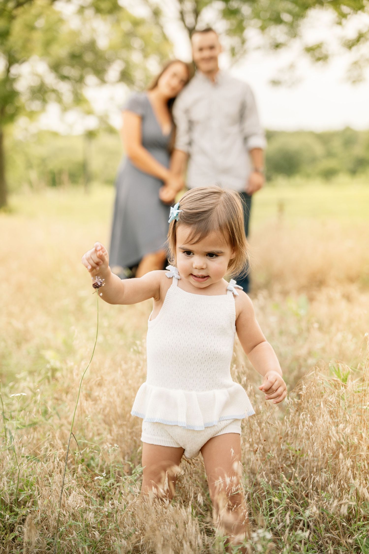 Toddler girl in white outfit walks through a sunny meadow while her parents embrace in the background.