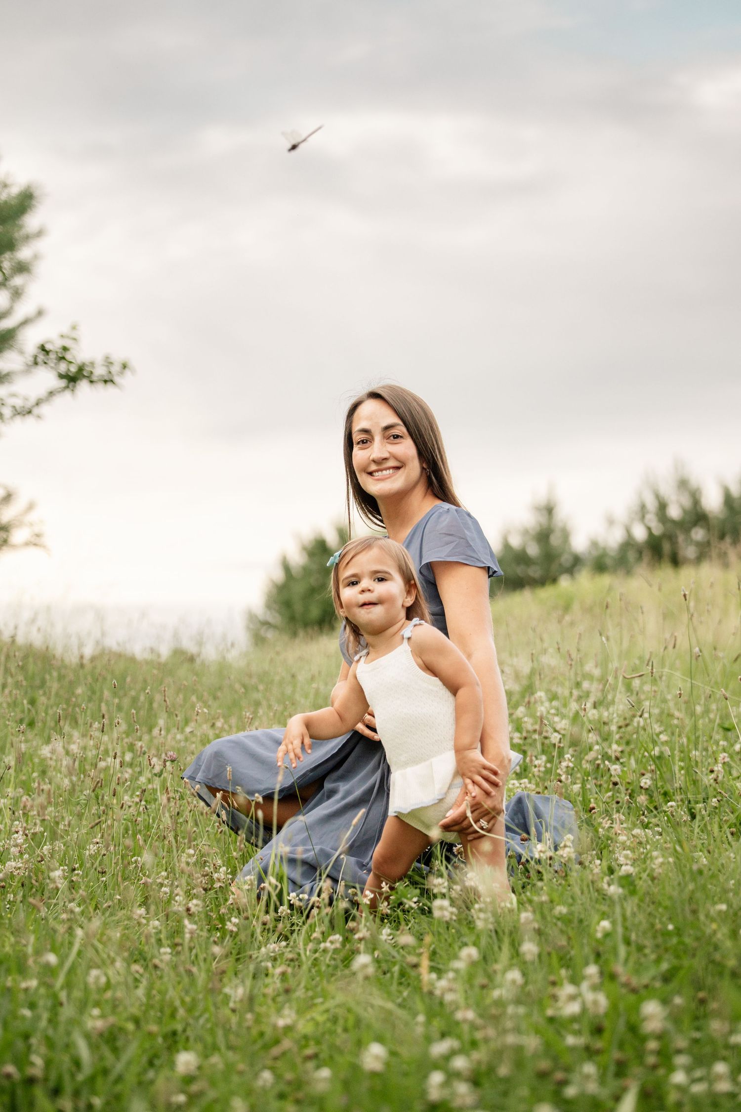 Smiling mother kneeling with toddler daughter in a clover-filled green meadow.