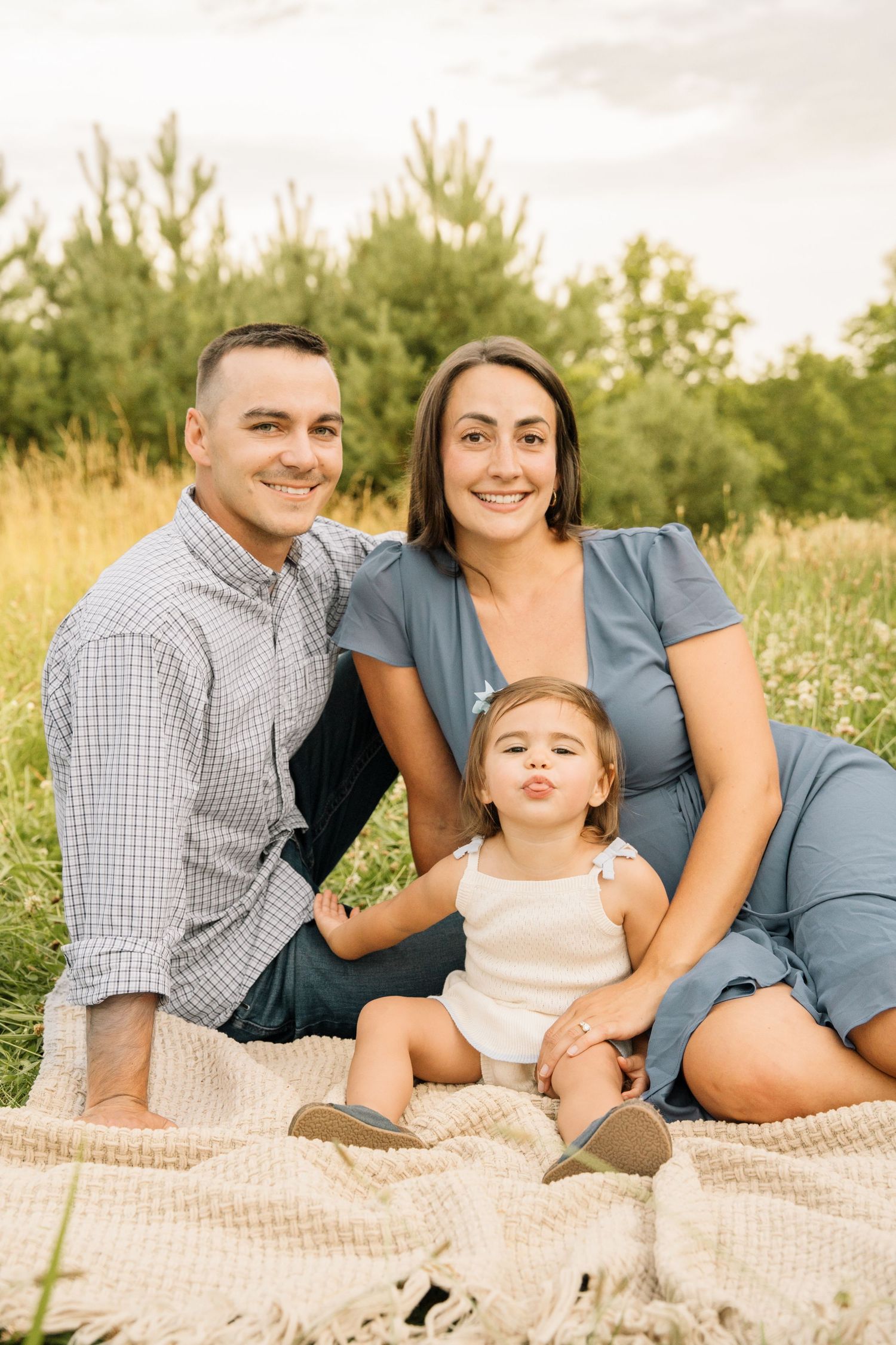 Smiling family of three posing on a blanket in a field, with their toddler making a funny face at the camera.