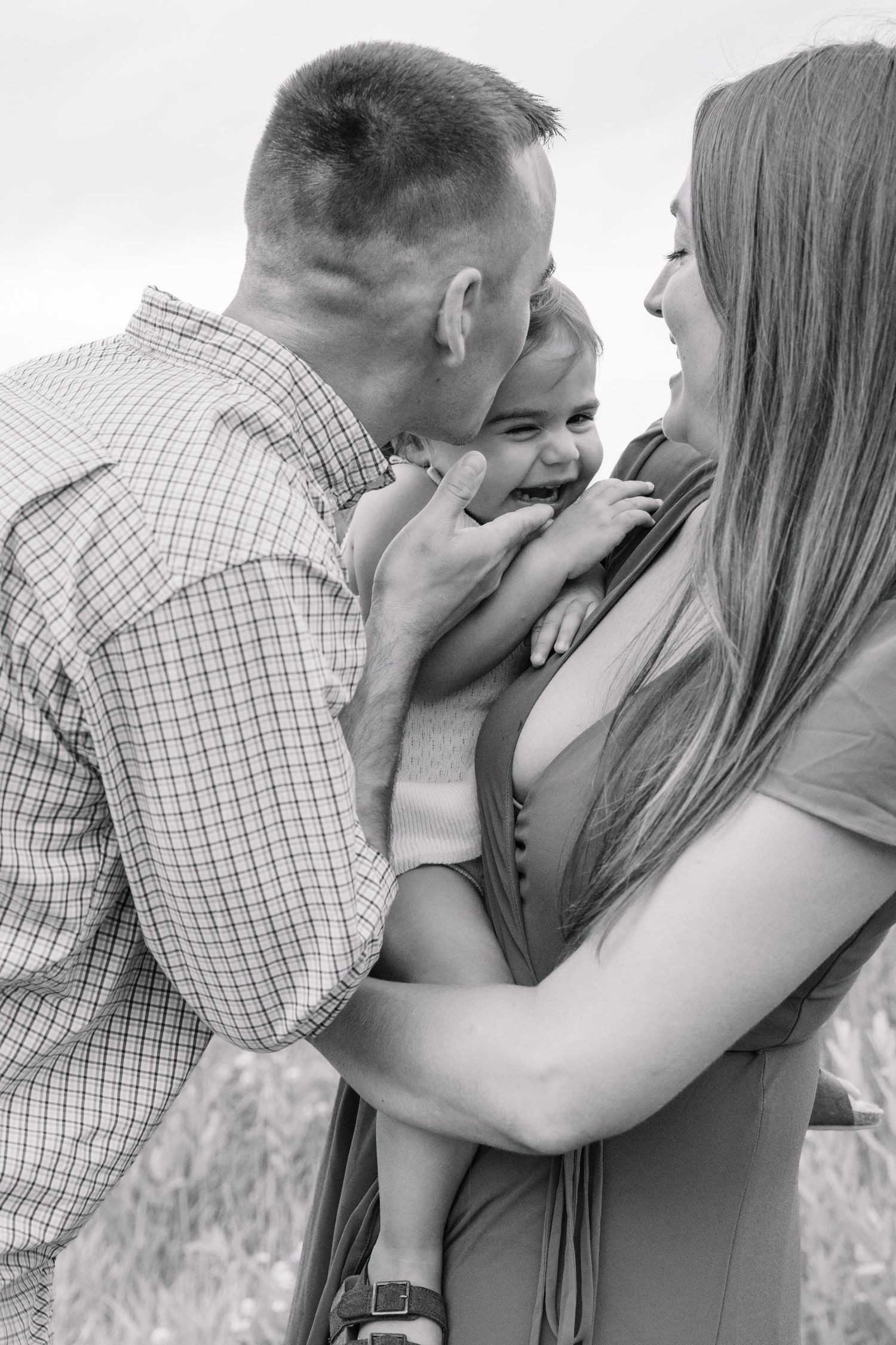 Black and white photo of happy parents kissing and cuddling their giggling baby outdoors in a field.