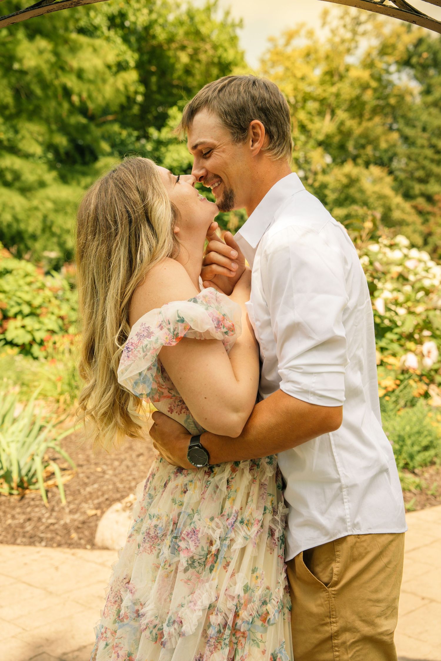 A couple embraces and smiles at each other in a lush garden, woman in floral dress, man in white shirt.