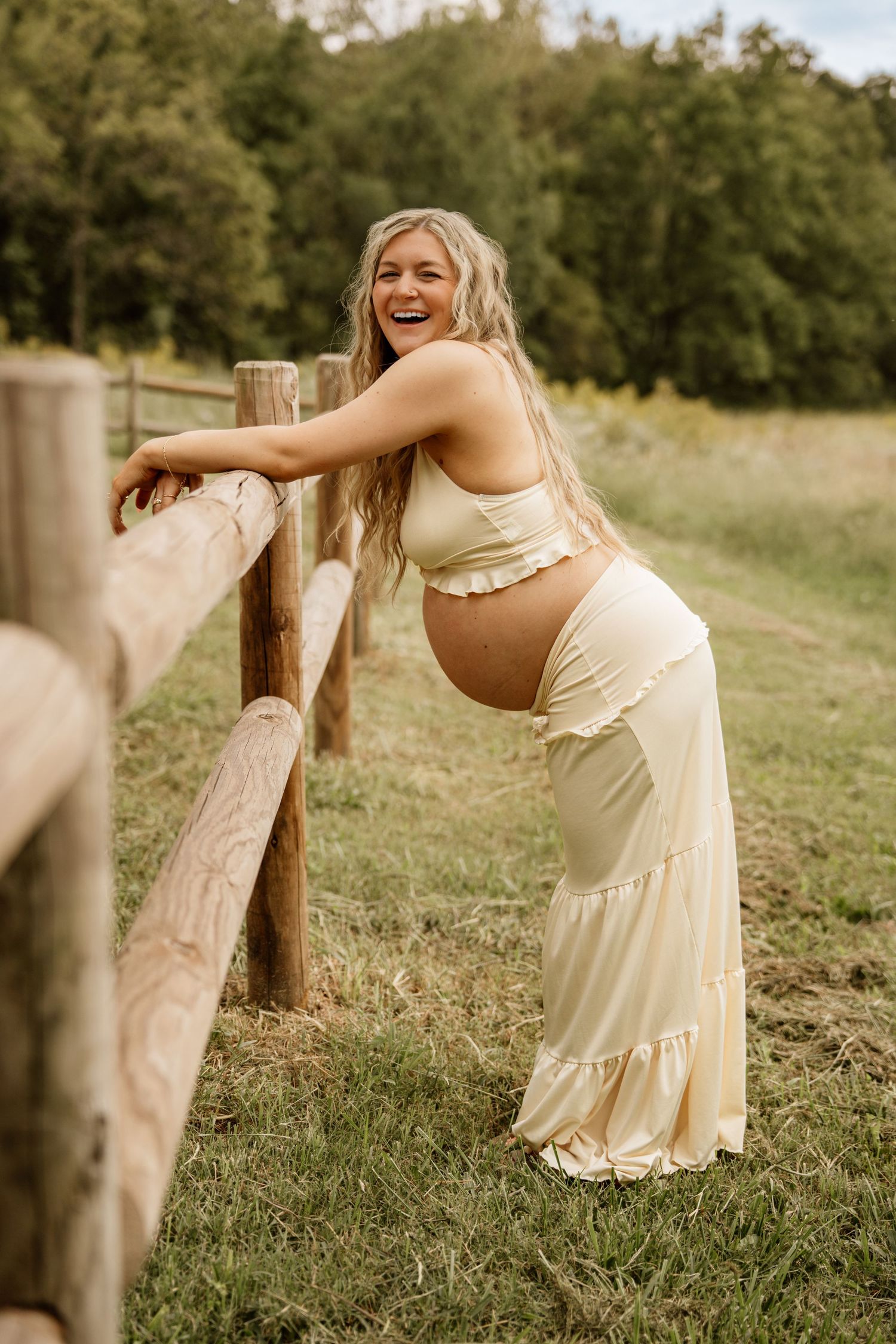 Laughing pregnant woman in cream crop top and maxi skirt leans on a rustic wooden fence.