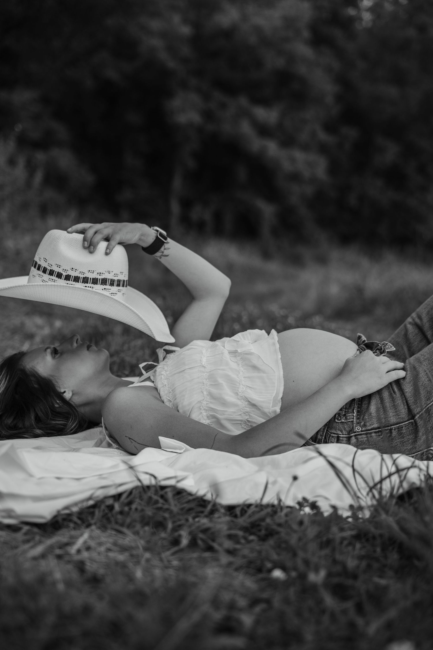 Black and white photo of pregnant woman lying on blanket in grass, holding cowboy hat over her face.