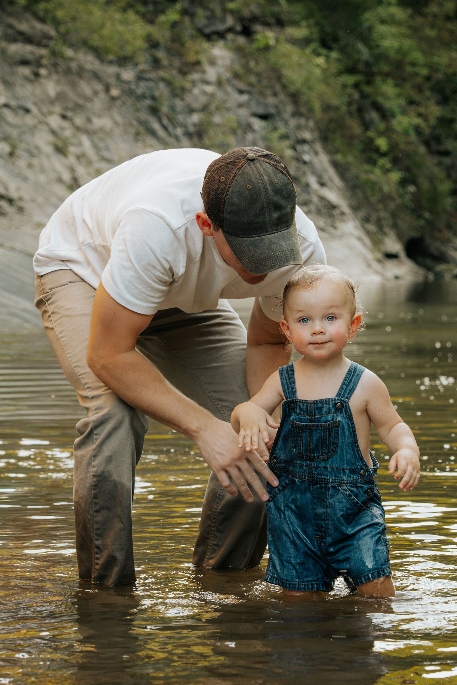 Father helping smiling toddler in denim overalls stand in a shallow river with rocky cliffs behind.