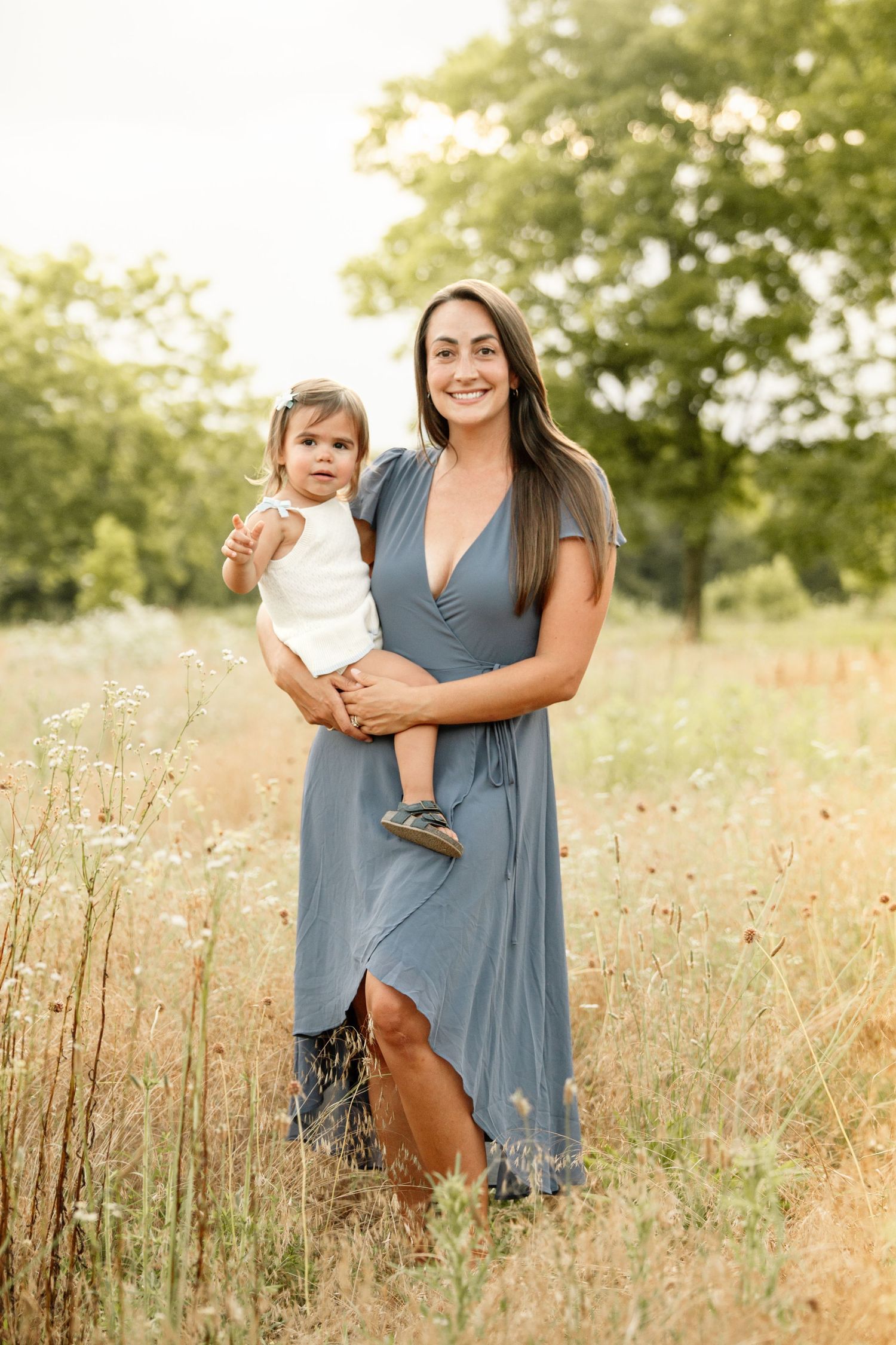 A smiling woman in a blue dress holds a baby in a sunlit wildflower field with green trees in the background.