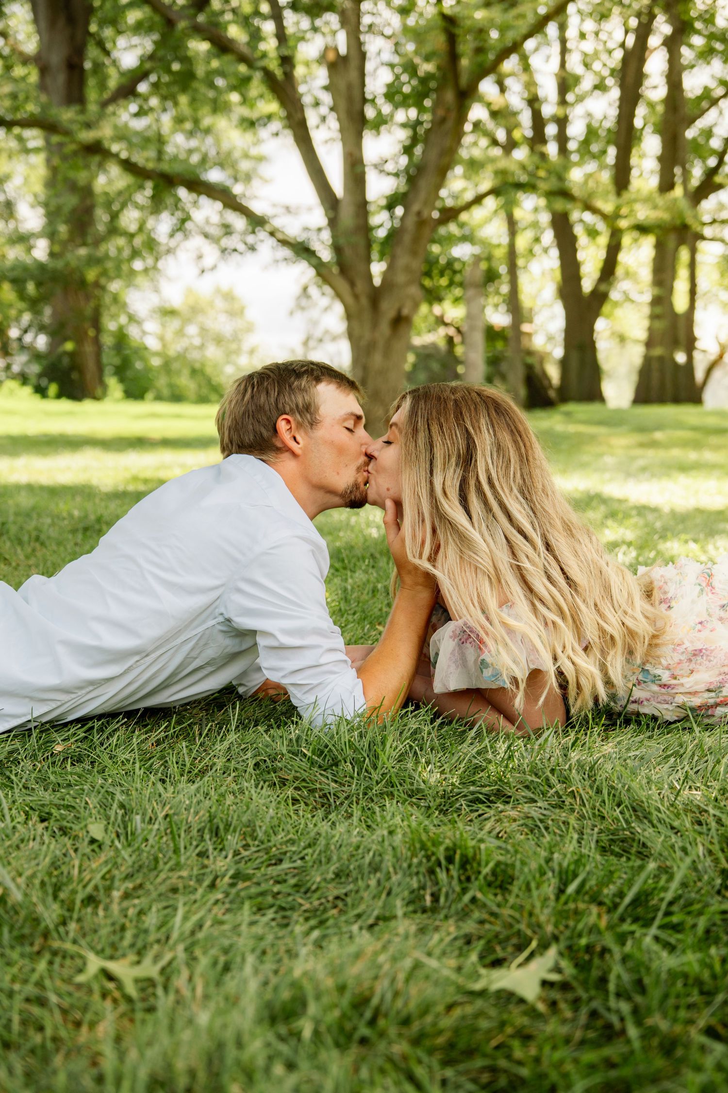 A couple lying on lush green grass in a park, sharing a romantic kiss beneath tall trees on a sunny day.