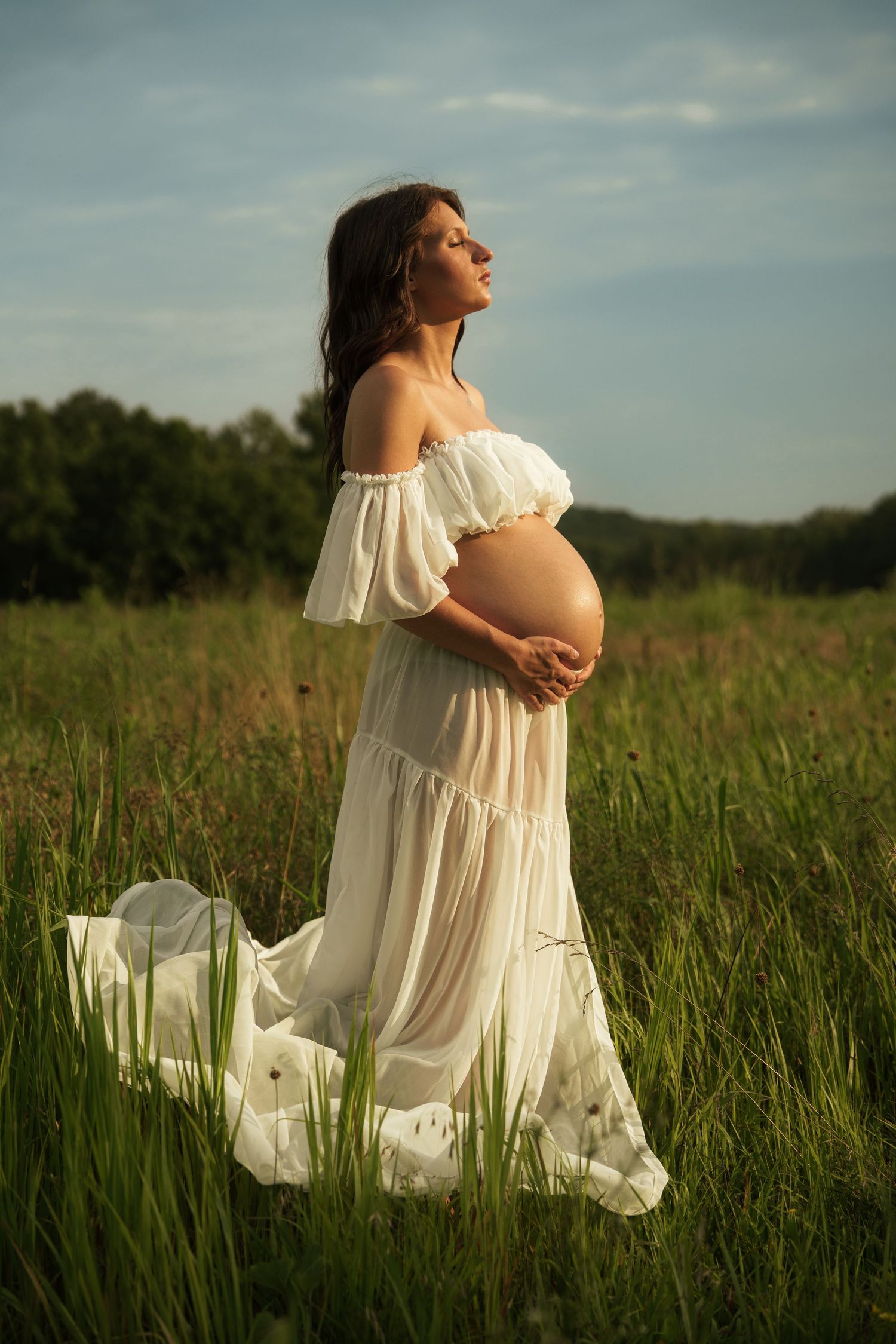 Pregnant woman in white off-shoulder dress standing in green field, cradling baby bump, golden hour sunlight.