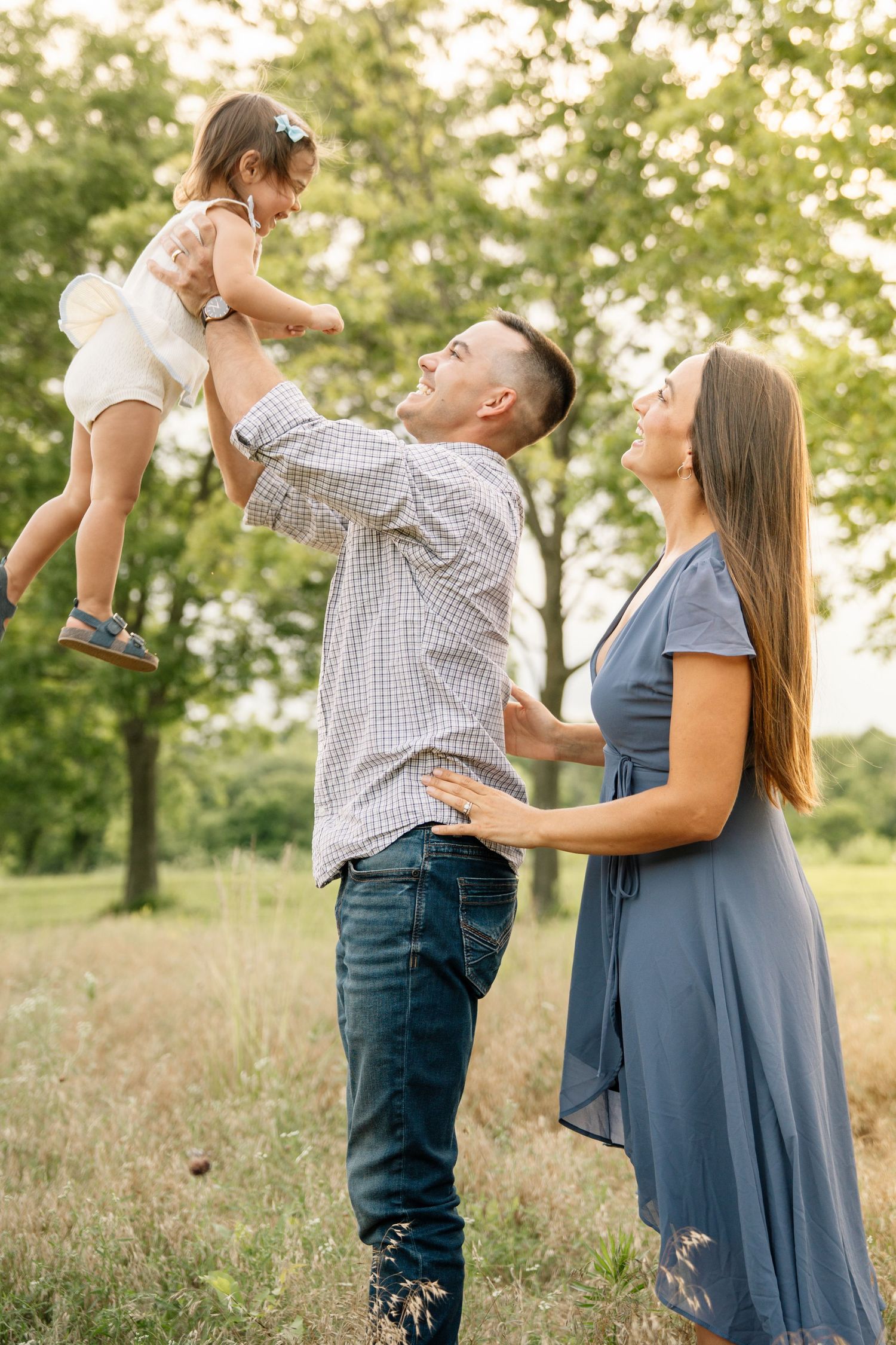 Happy family playing outdoors, father lifting toddler girl in white dress while mother smiles in blue dress, green park.
