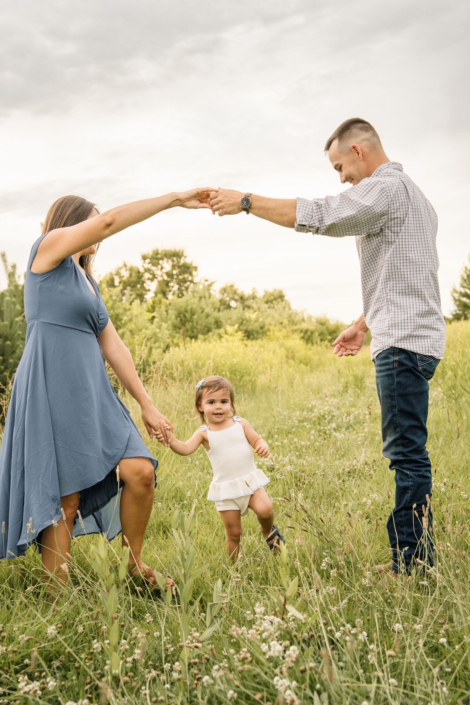 Parents holding hands above toddler daughter dancing in a wildflower meadow on a cloudy day.