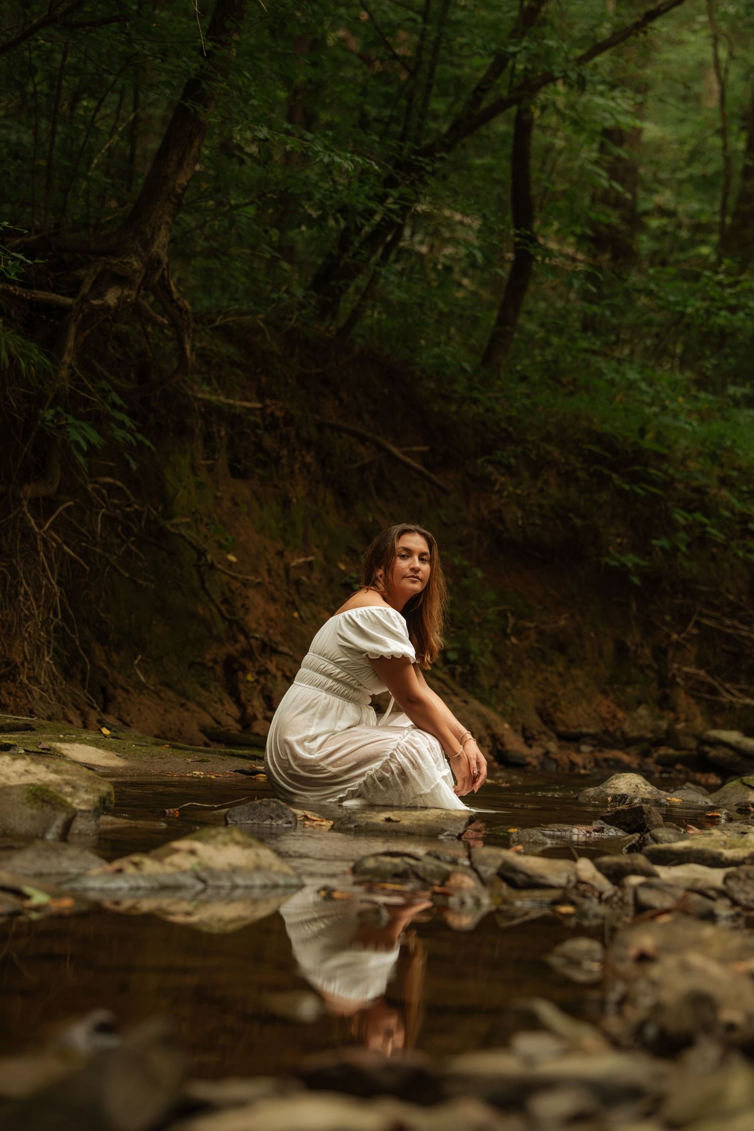 Woman in white dress sitting on rocks by a forest stream, her reflection visible in the calm water below.