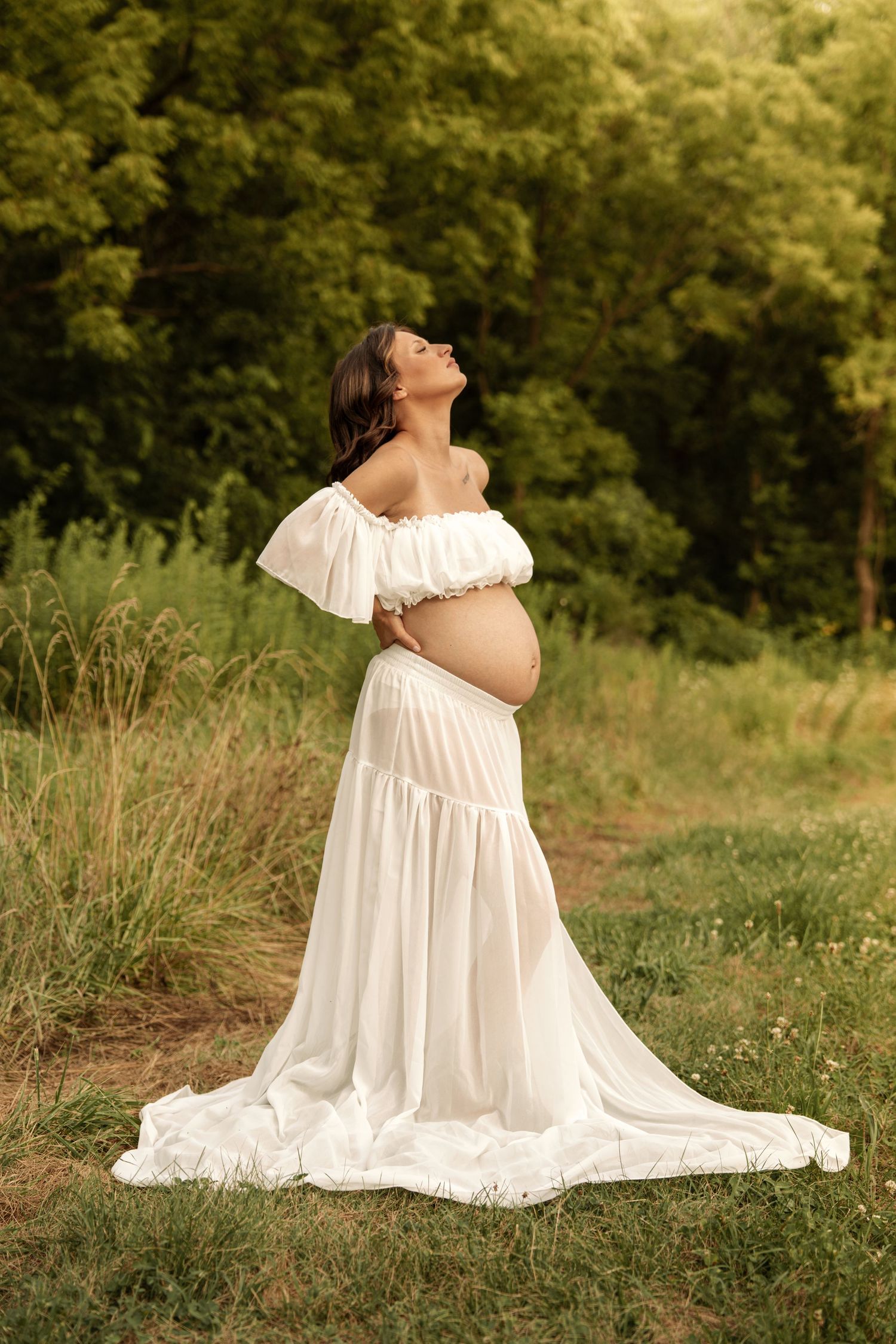 Pregnant woman in a flowing white dress tilting her head back gracefully in a lush green field.