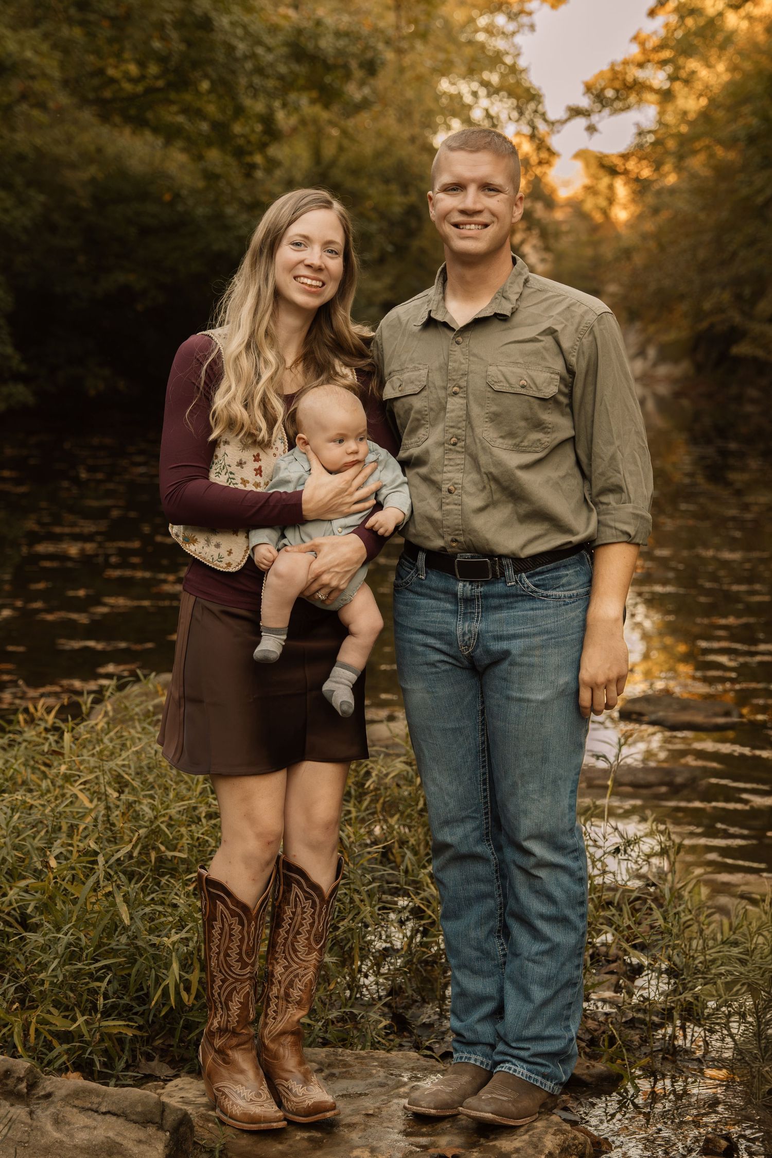 Young family with a baby poses outdoors near a creek surrounded by autumn foliage in warm golden light.