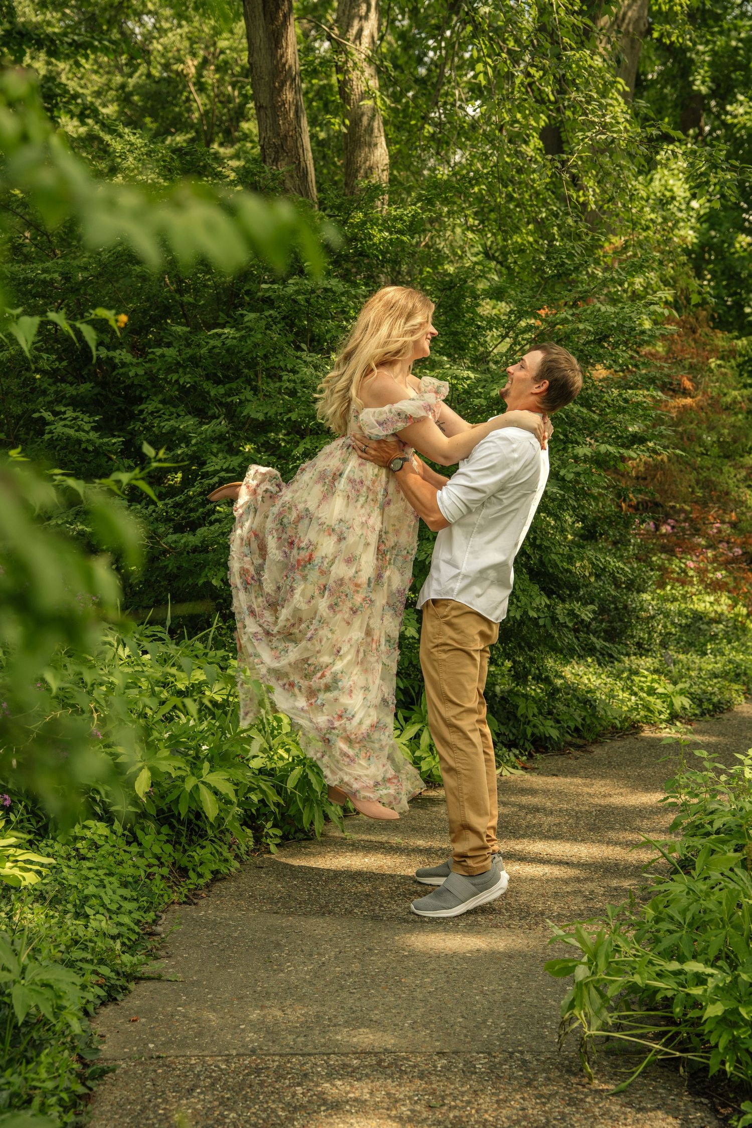 A man lifts a woman in a floral dress on a lush green garden path.