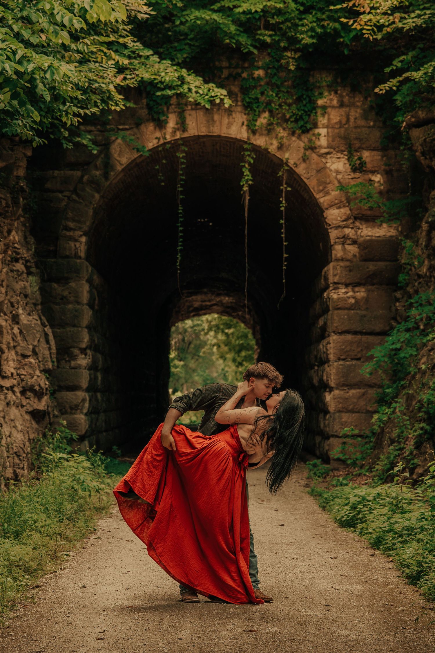 Couple sharing a romantic dip kiss in front of a stone arch tunnel, woman in flowing red gown.