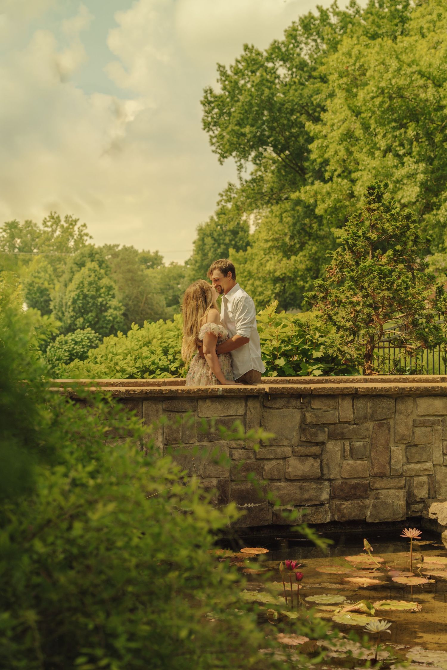 Romantic couple sharing an intimate moment on a stone bridge over a lily pond in a lush green garden.