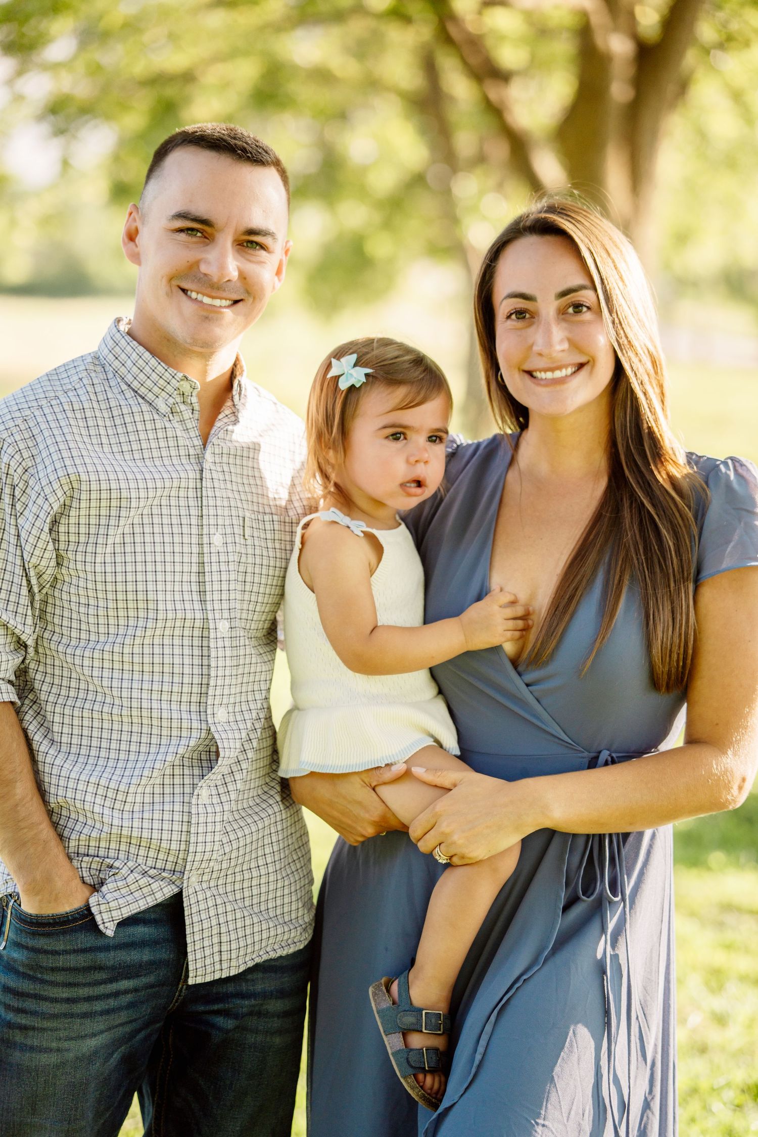 Happy family portrait with smiling parents holding toddler girl outdoors in sunny green park.