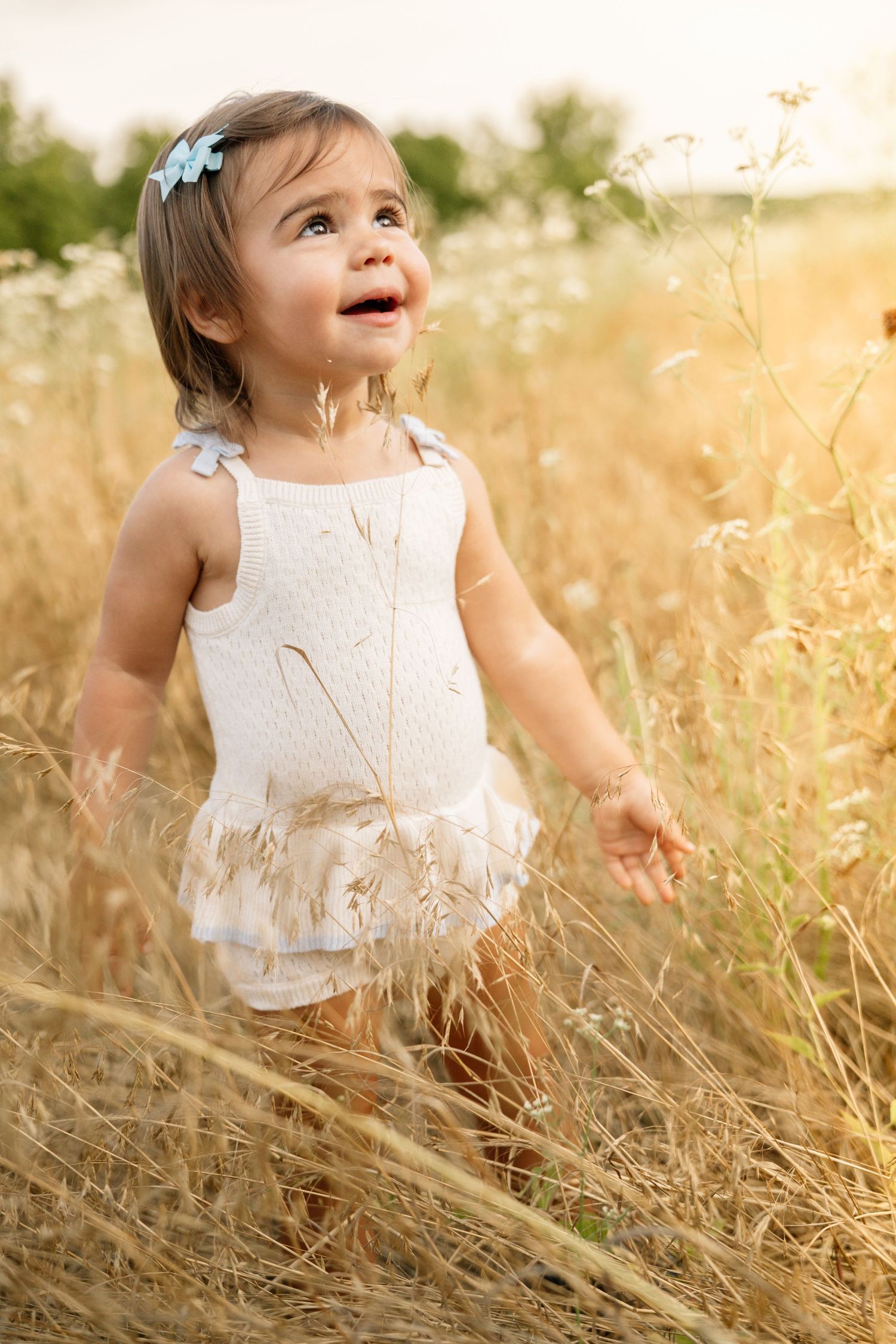 Smiling toddler girl in white dress standing in golden field of tall grass and wildflowers.
