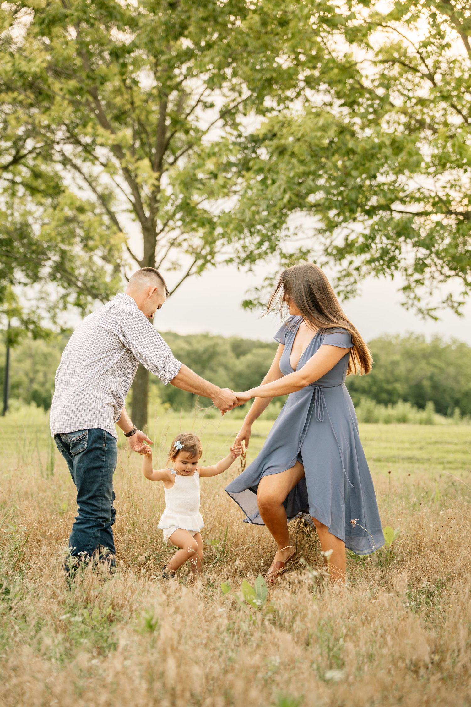 A happy family of three plays together in a sunny meadow, with parents holding their toddler daughter's hands outdoors.