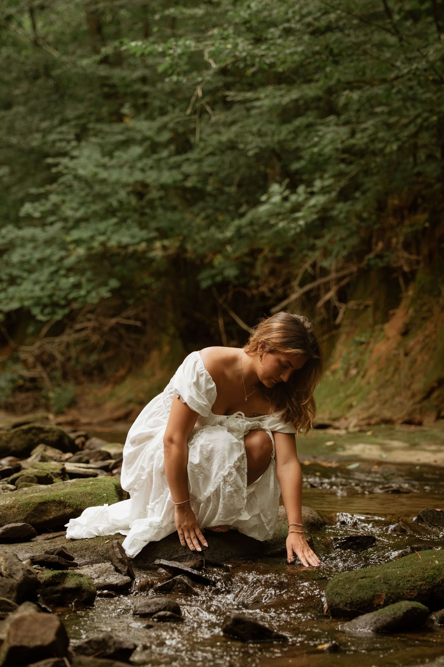 Woman in white off-shoulder dress touching a forest stream while crouching on rocks.