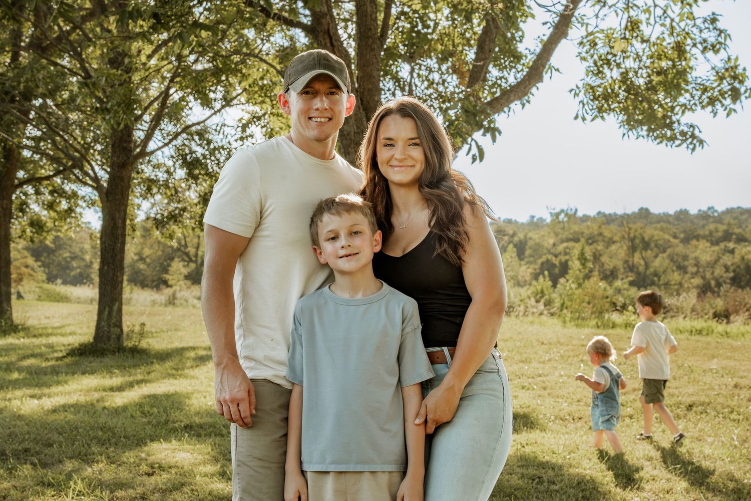 Family of three posing outdoors in a green park setting with trees, smiling warmly on a sunny day.