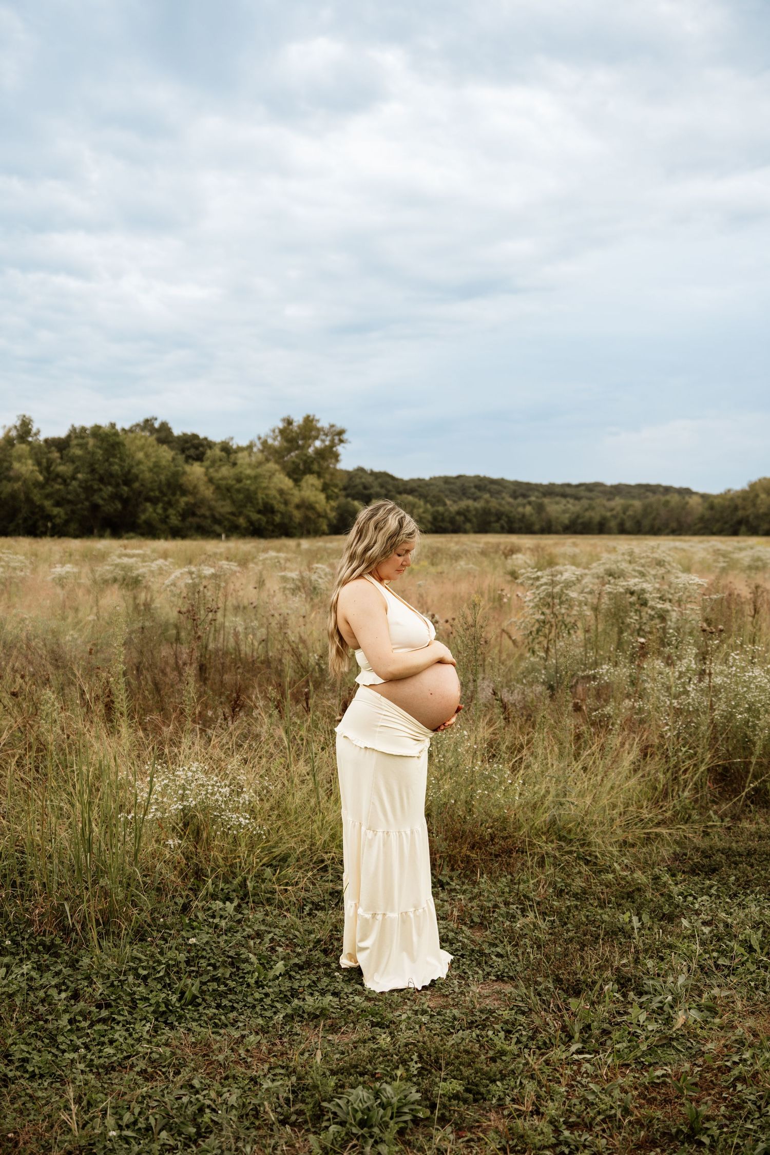 Pregnant woman in white maternity dress stands sideways in an open field under cloudy skies.