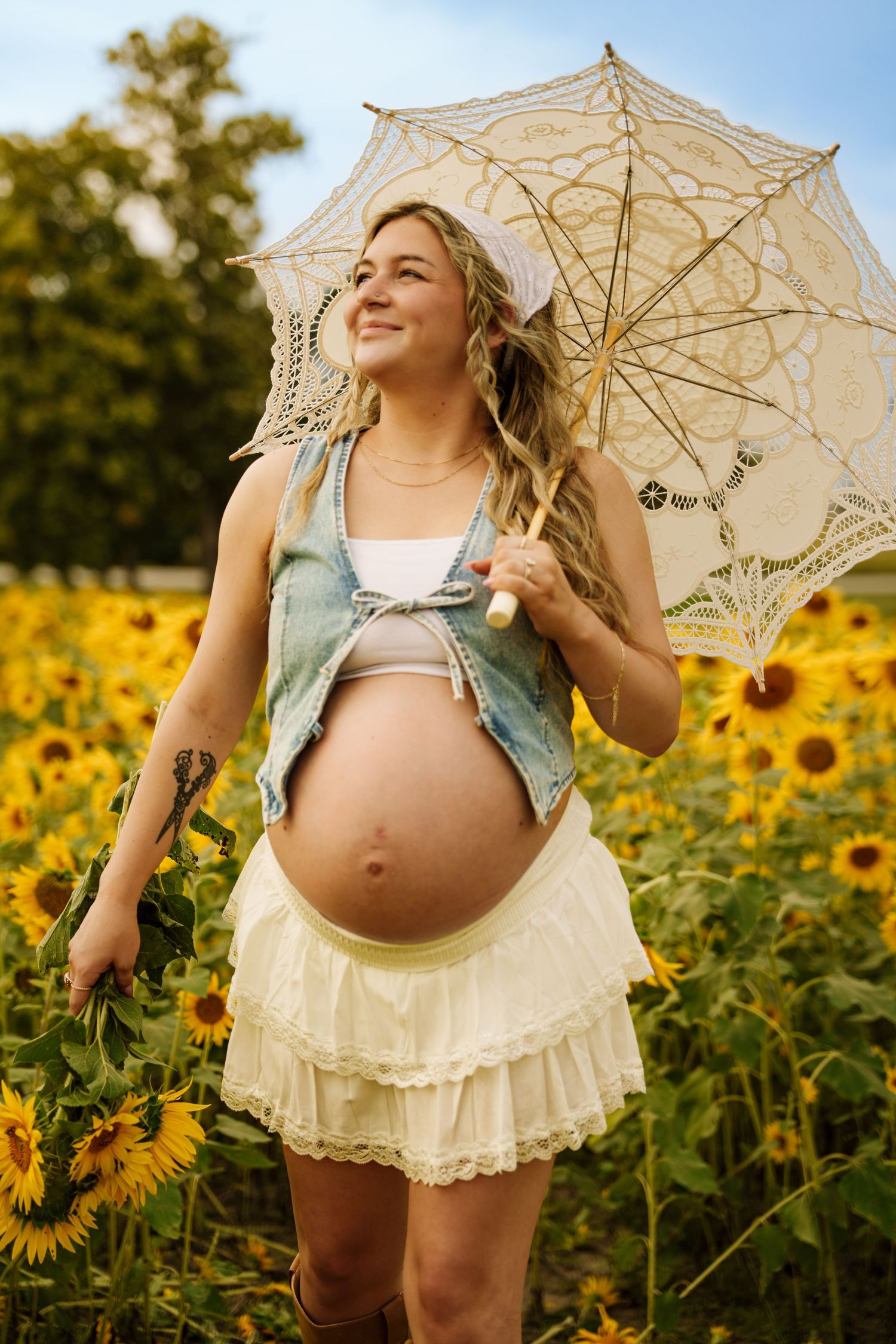 Smiling pregnant woman in sunflower field holding lace parasol, wearing denim vest and cream ruffled skirt.