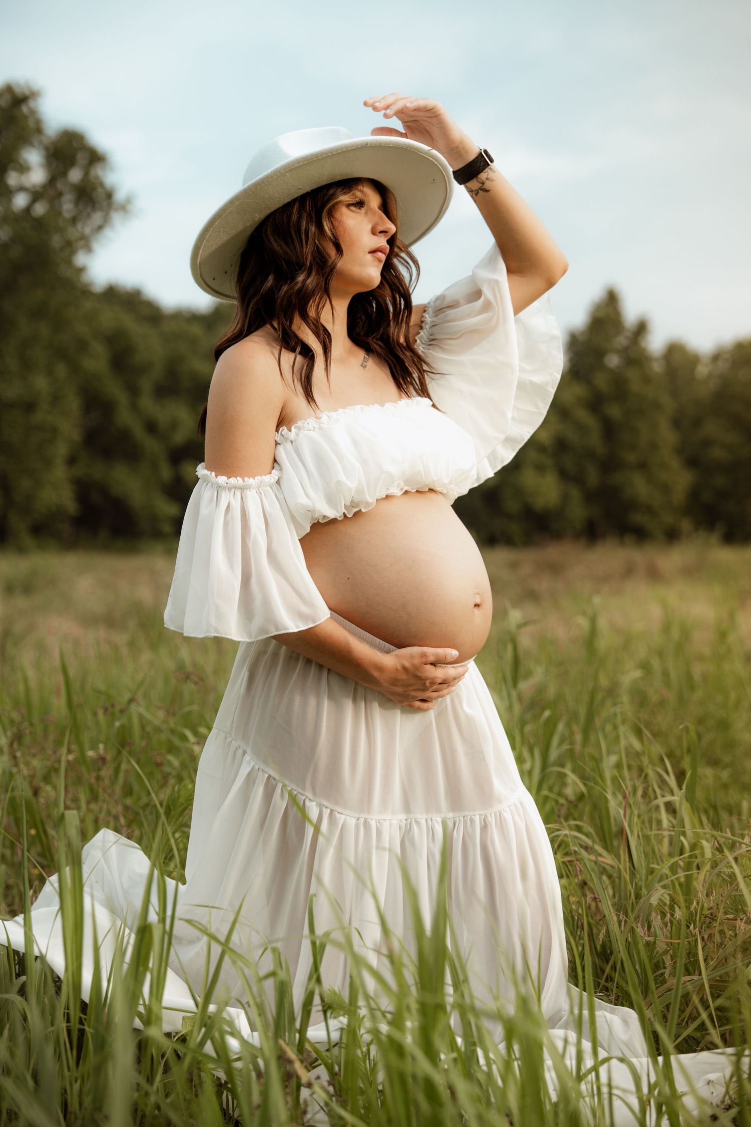 Pregnant woman in white dress and hat cradling her bump while kneeling in a green field.