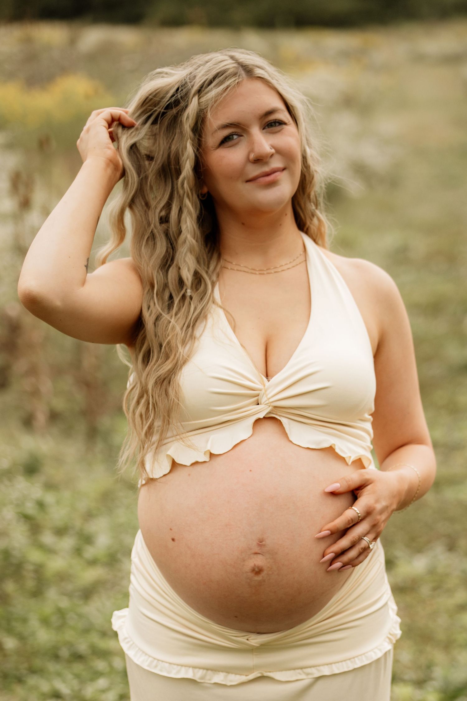 Smiling pregnant woman with wavy blonde hair in cream halter top cradling baby bump outdoors.