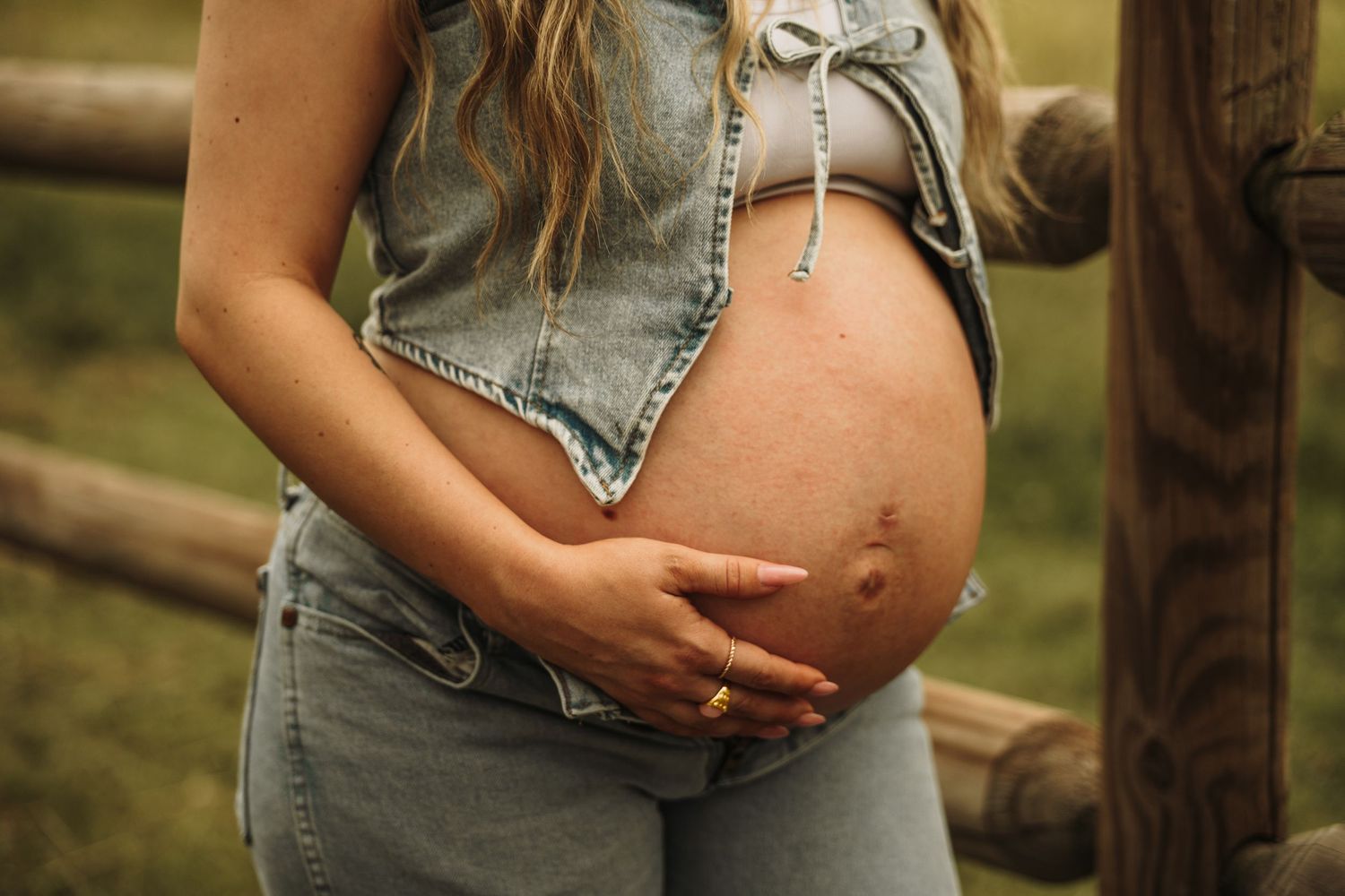 Pregnant woman in denim vest and jeans cradling bare baby bump outdoors near rustic wooden fence.