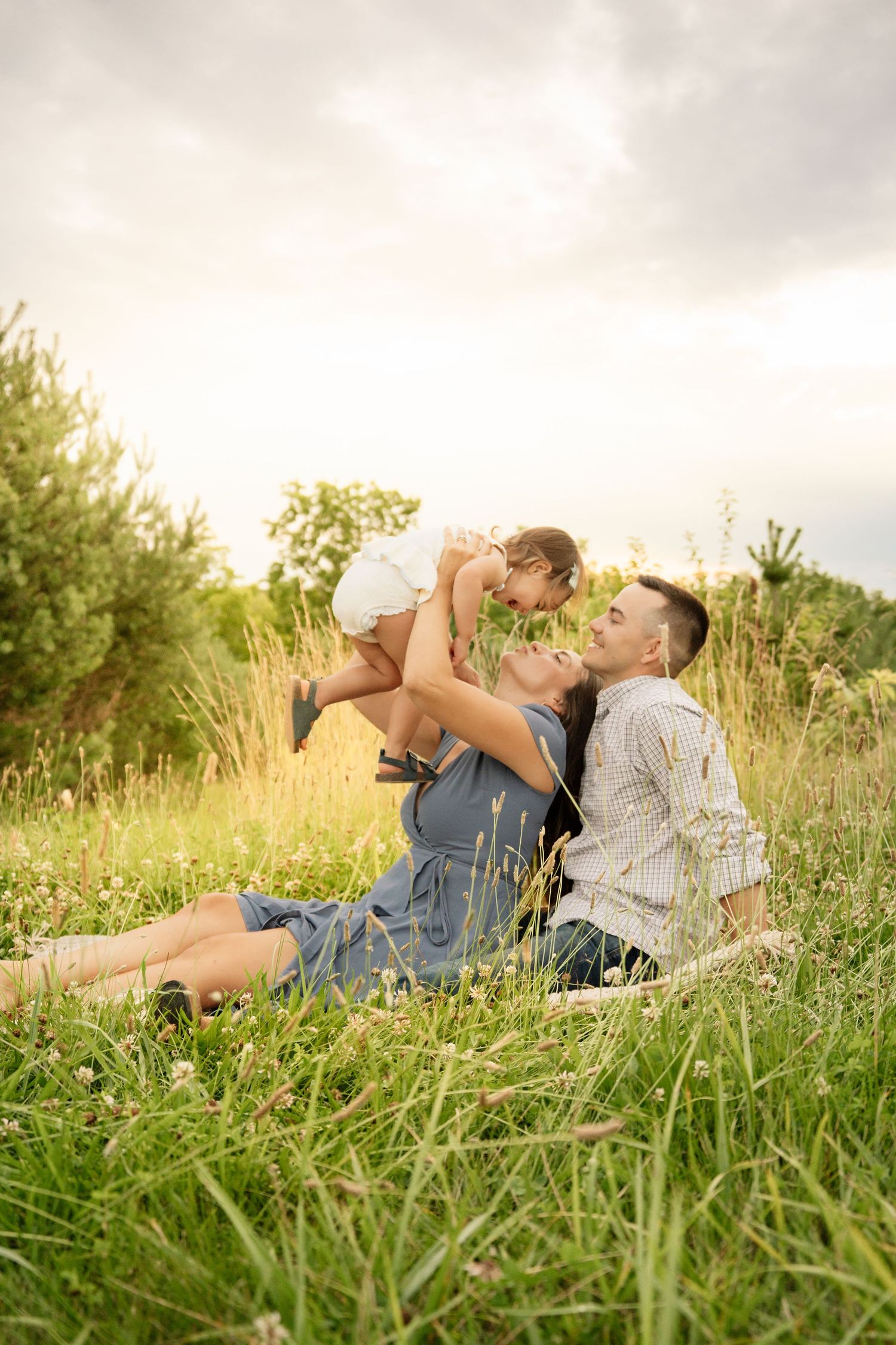 Happy family sitting in a sunny meadow, parents lifting and kissing their toddler outdoors.