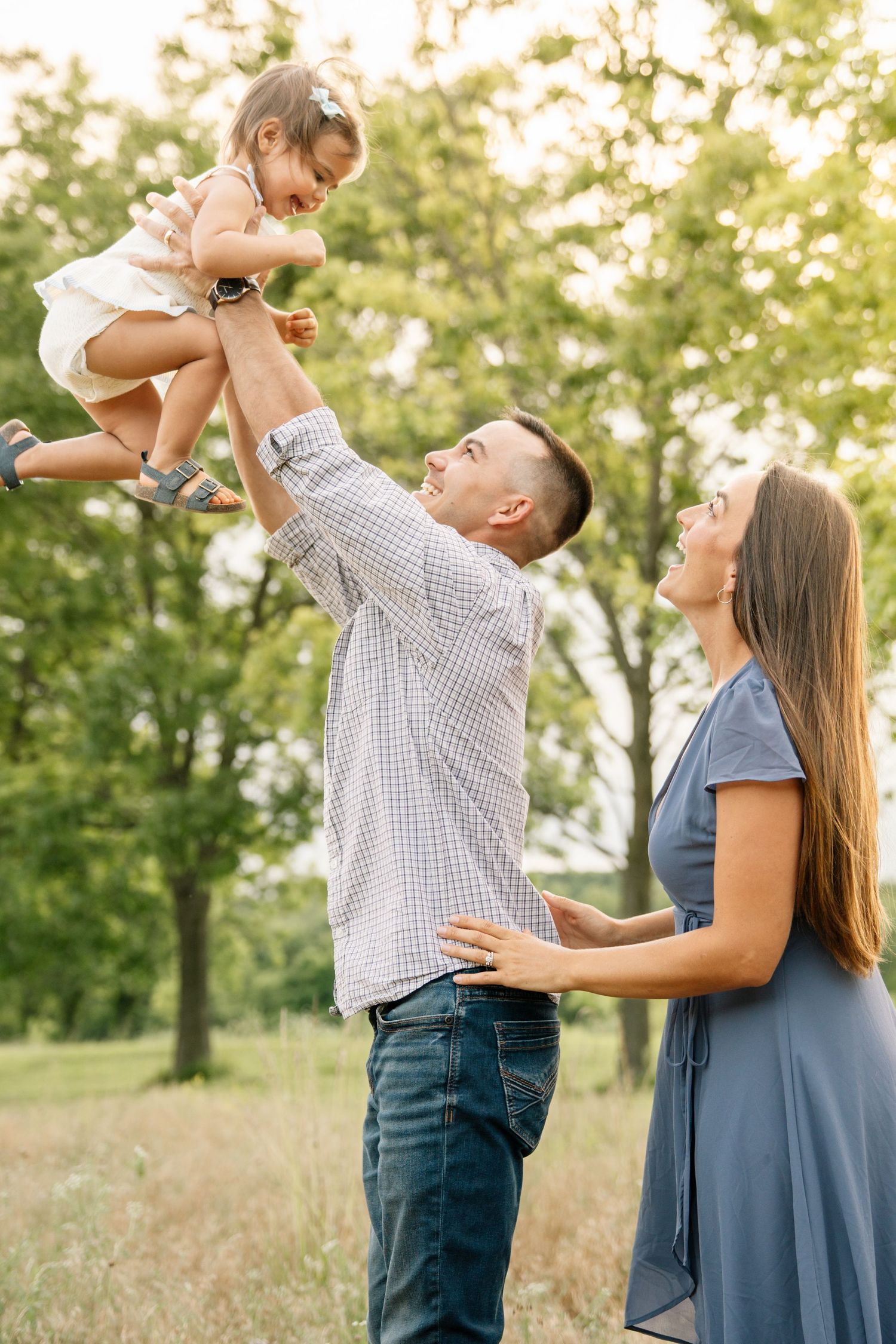 Happy family playing outdoors, father lifting toddler girl in air while mother watches and smiles in green park.
