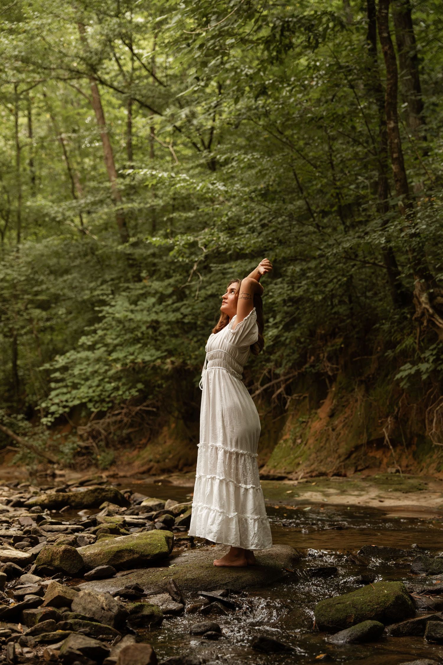 Woman in white dress stands on rocks by forest stream, arms raised skyward, embracing nature.