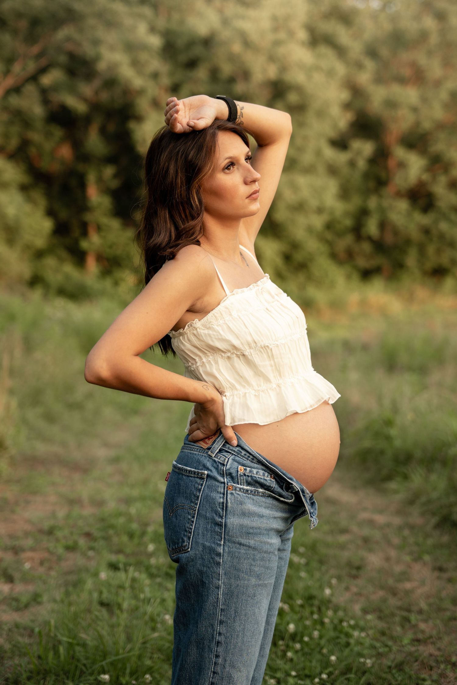 Pregnant woman in white crop top and jeans posing in a sunny green field.