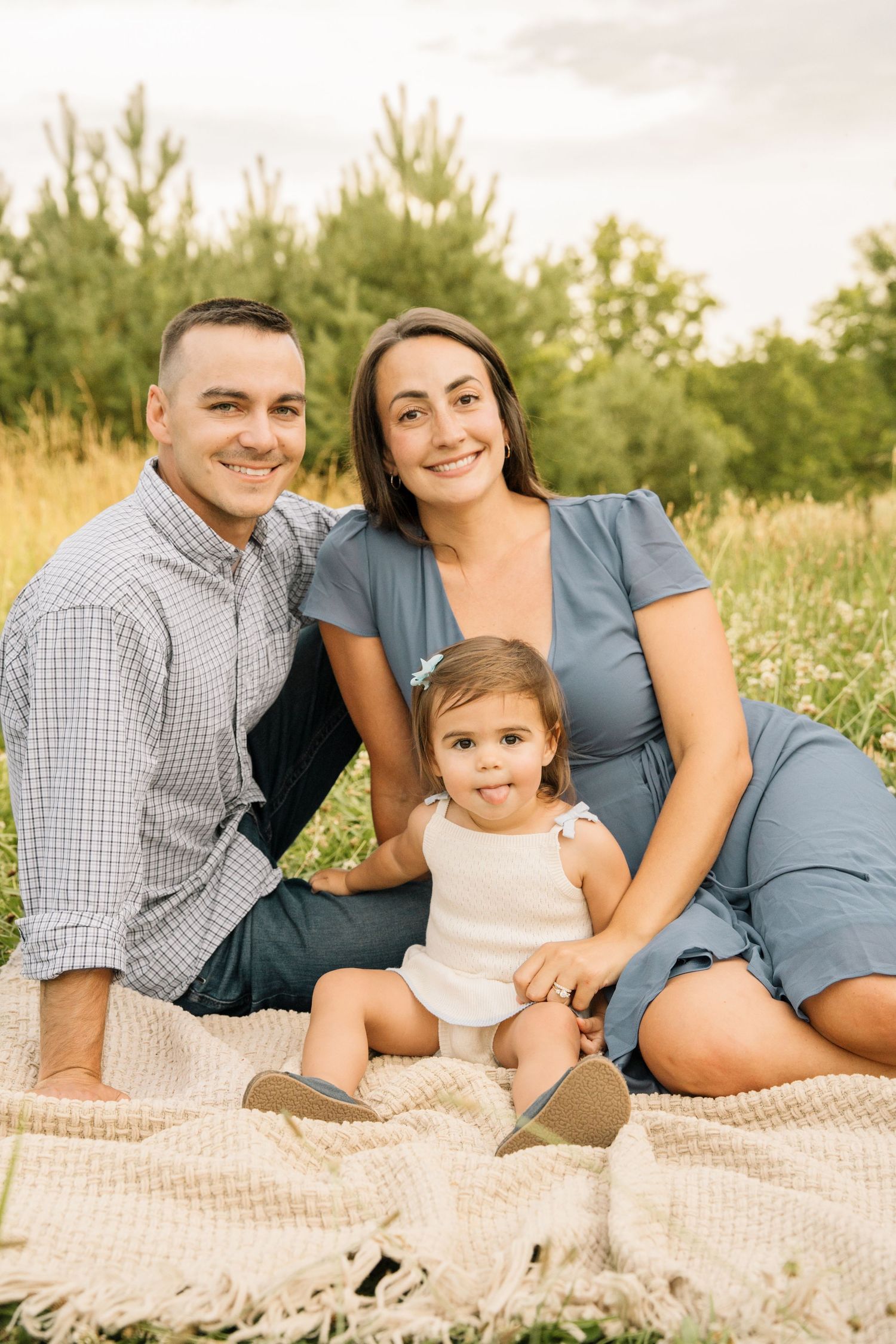 A happy family of three sits on a blanket in a green field, with parents smiling and a toddler in white romper.