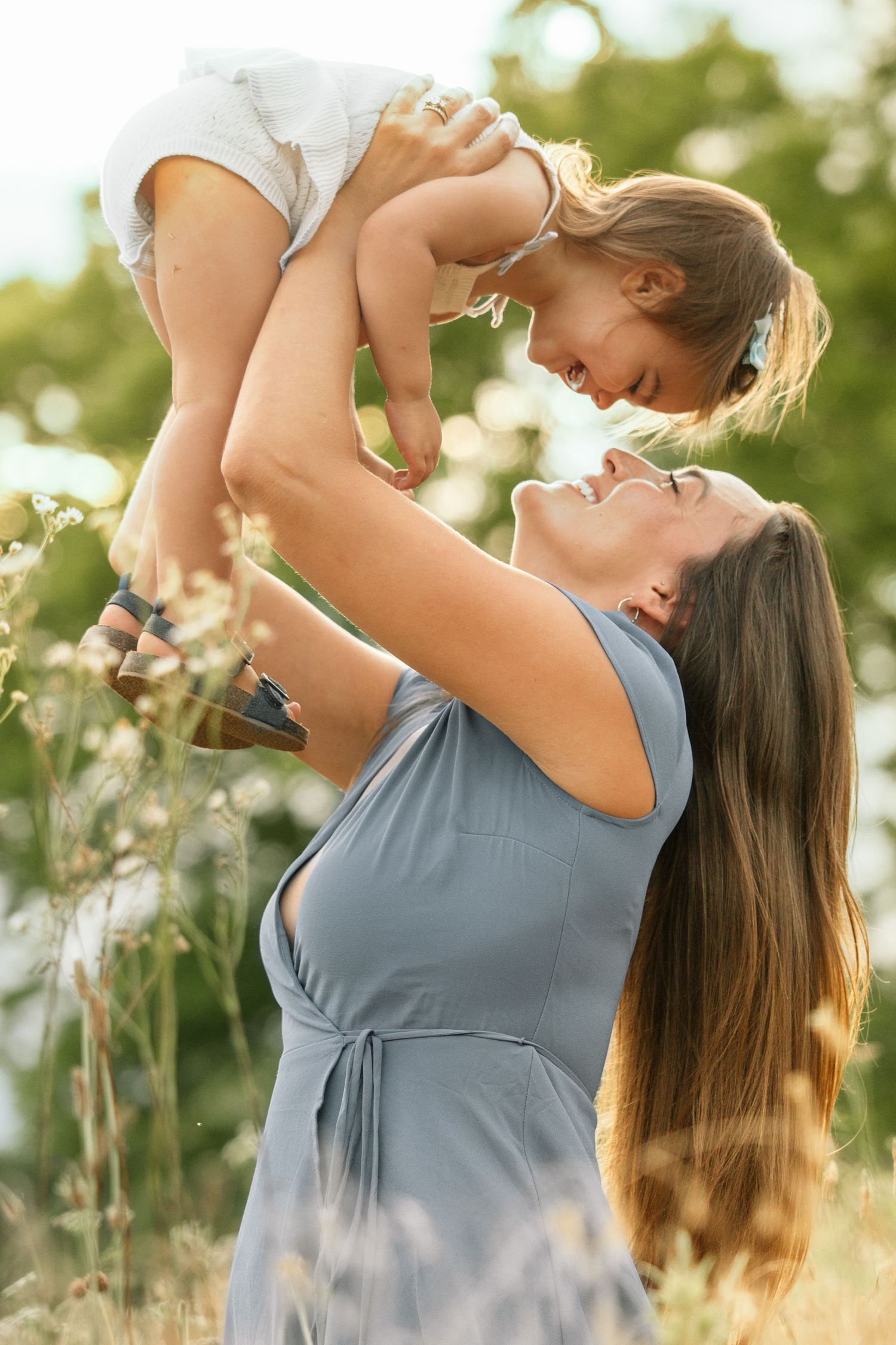Mother in blue dress raising smiling toddler girl overhead in sunny wildflower meadow.