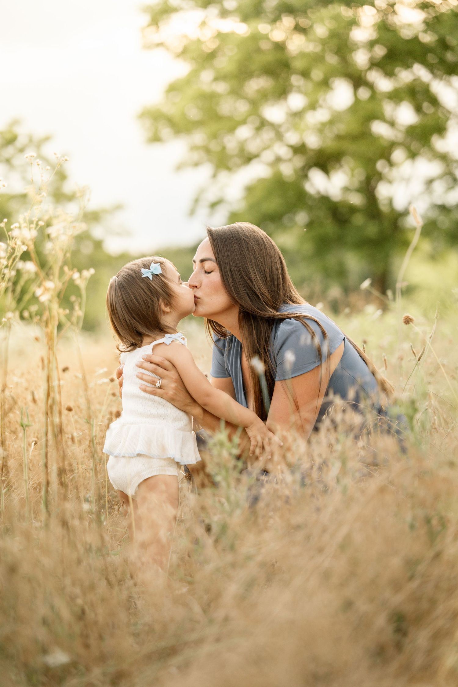 Mother kissing young toddler daughter in sun-drenched golden grass field outdoors.